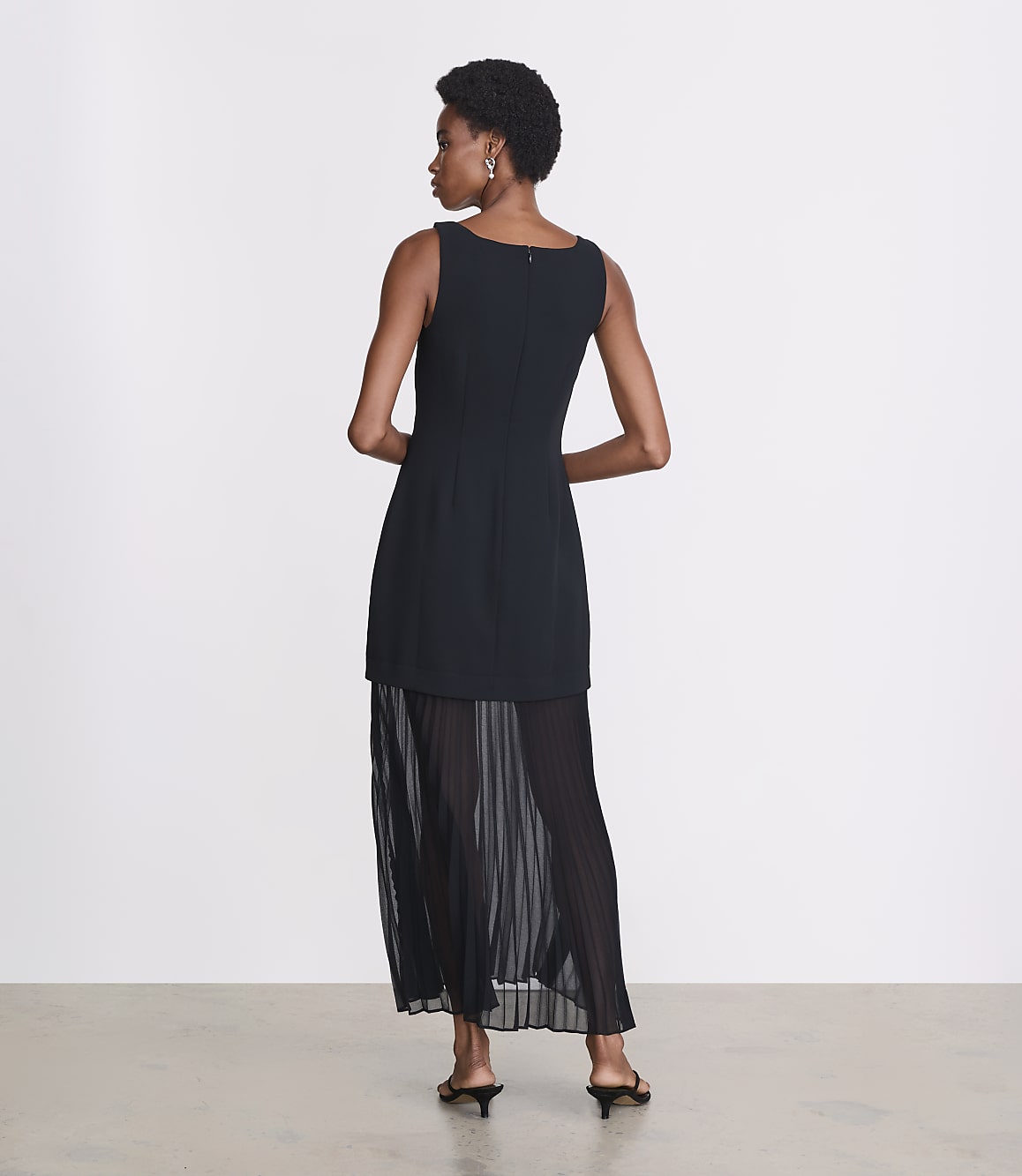 A woman in a black dress with a pleated skirt stands against a plain white background.