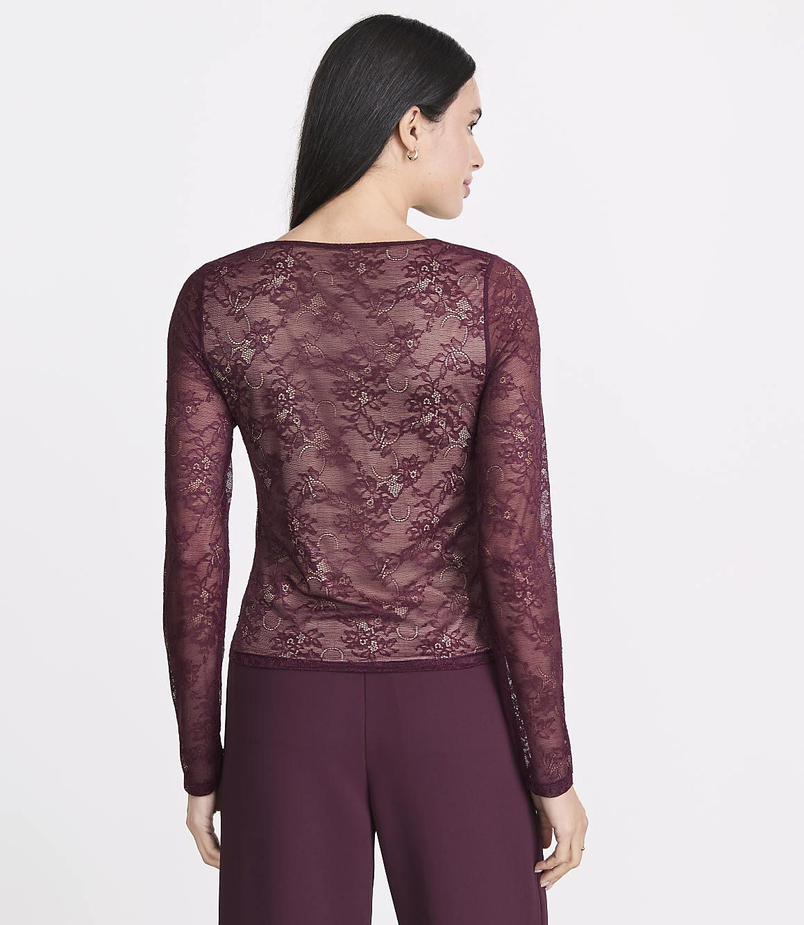 A woman in a sheer burgundy lace top and matching pants stands against a plain white background.