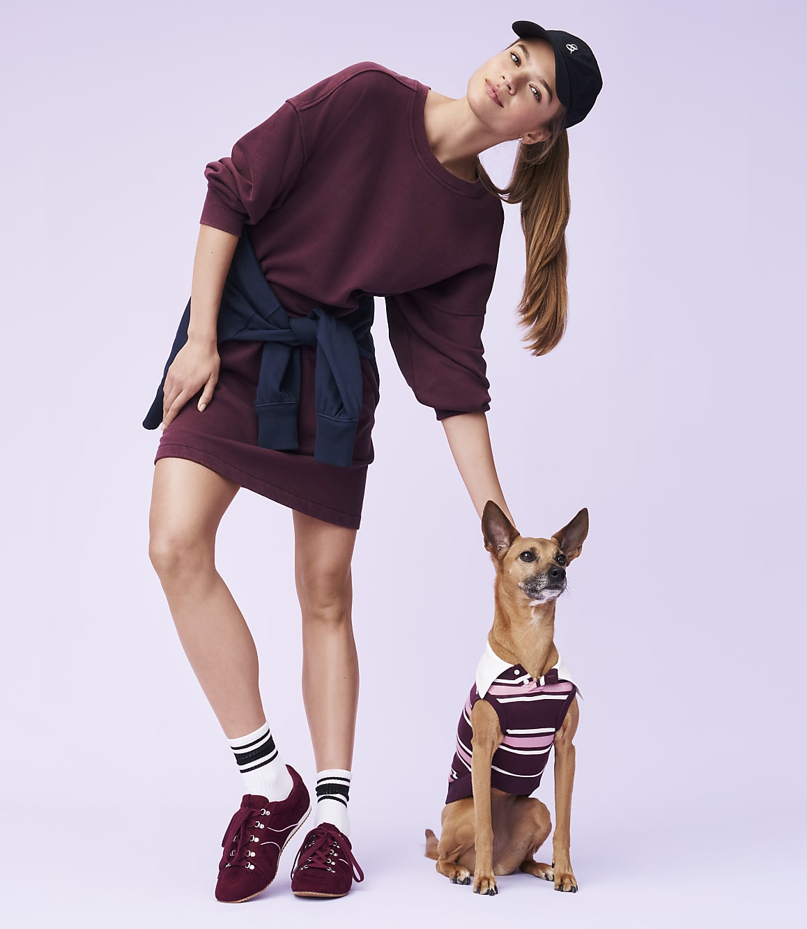 A young woman in a maroon dress and baseball cap poses with a small dog wearing a striped shirt against a light purple background.