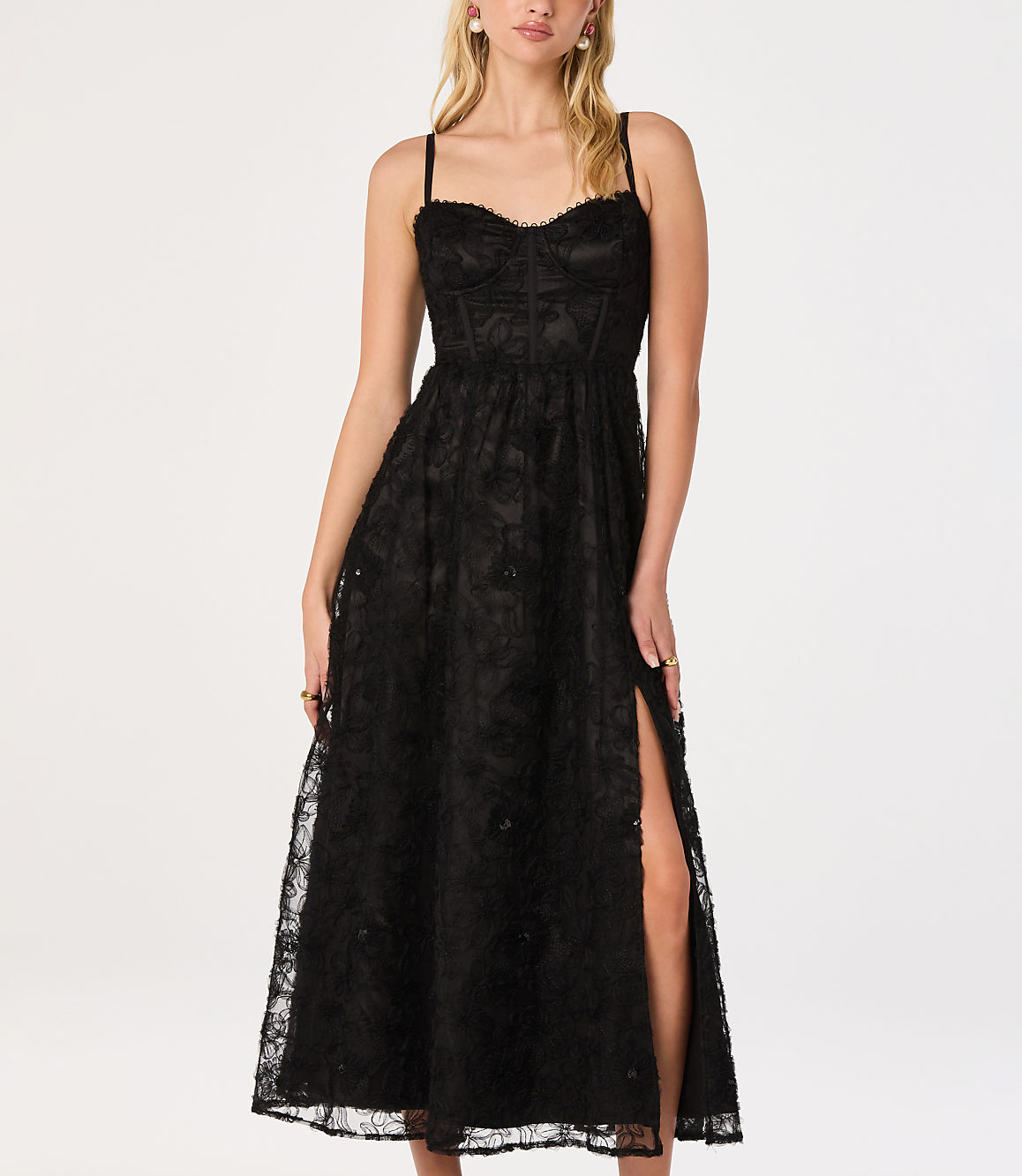 A woman in a black lace dress with a thigh-high slit stands against a plain white background.