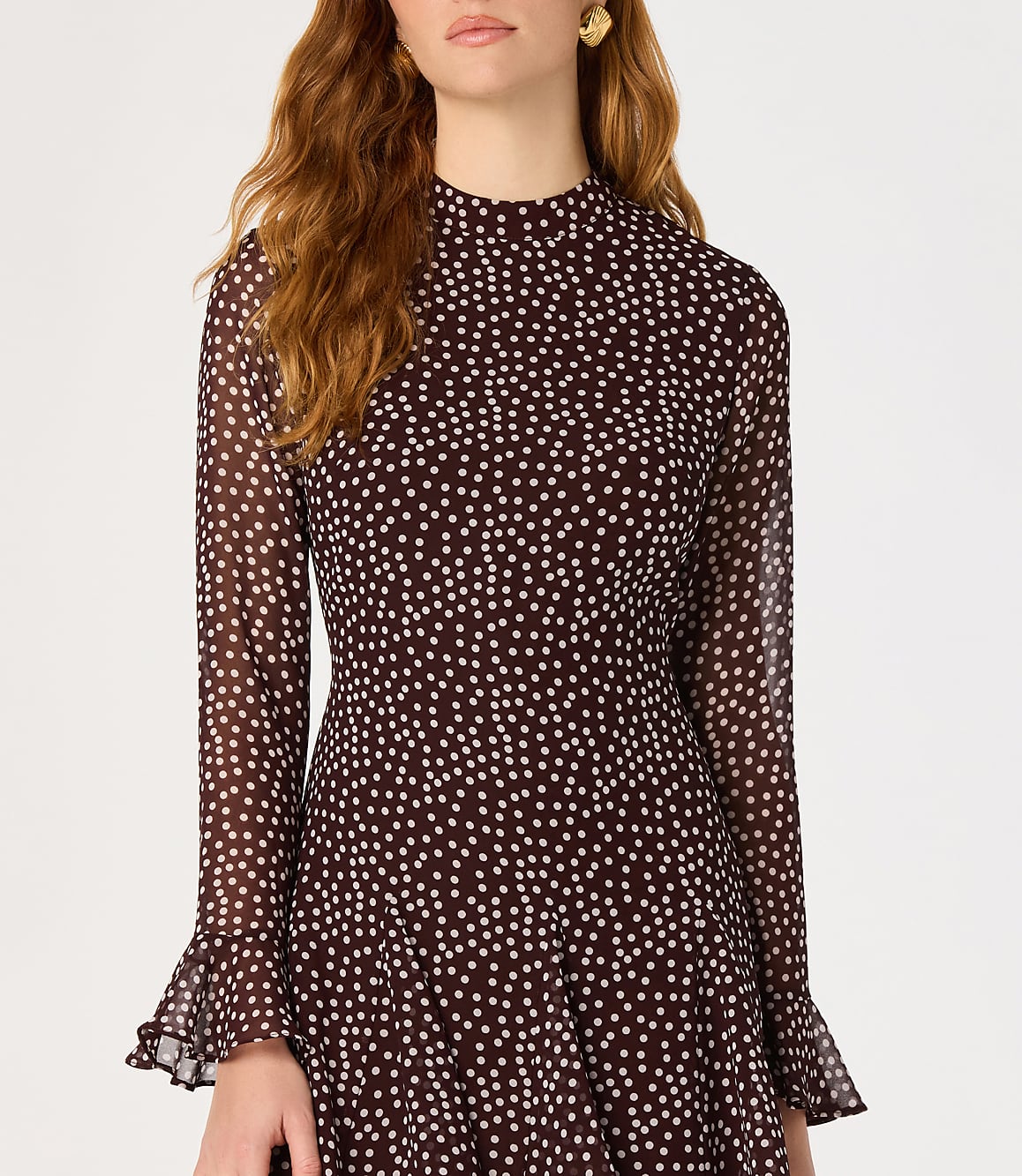 A woman in a dark, polka-dotted dress with bell sleeves stands against a plain white background.