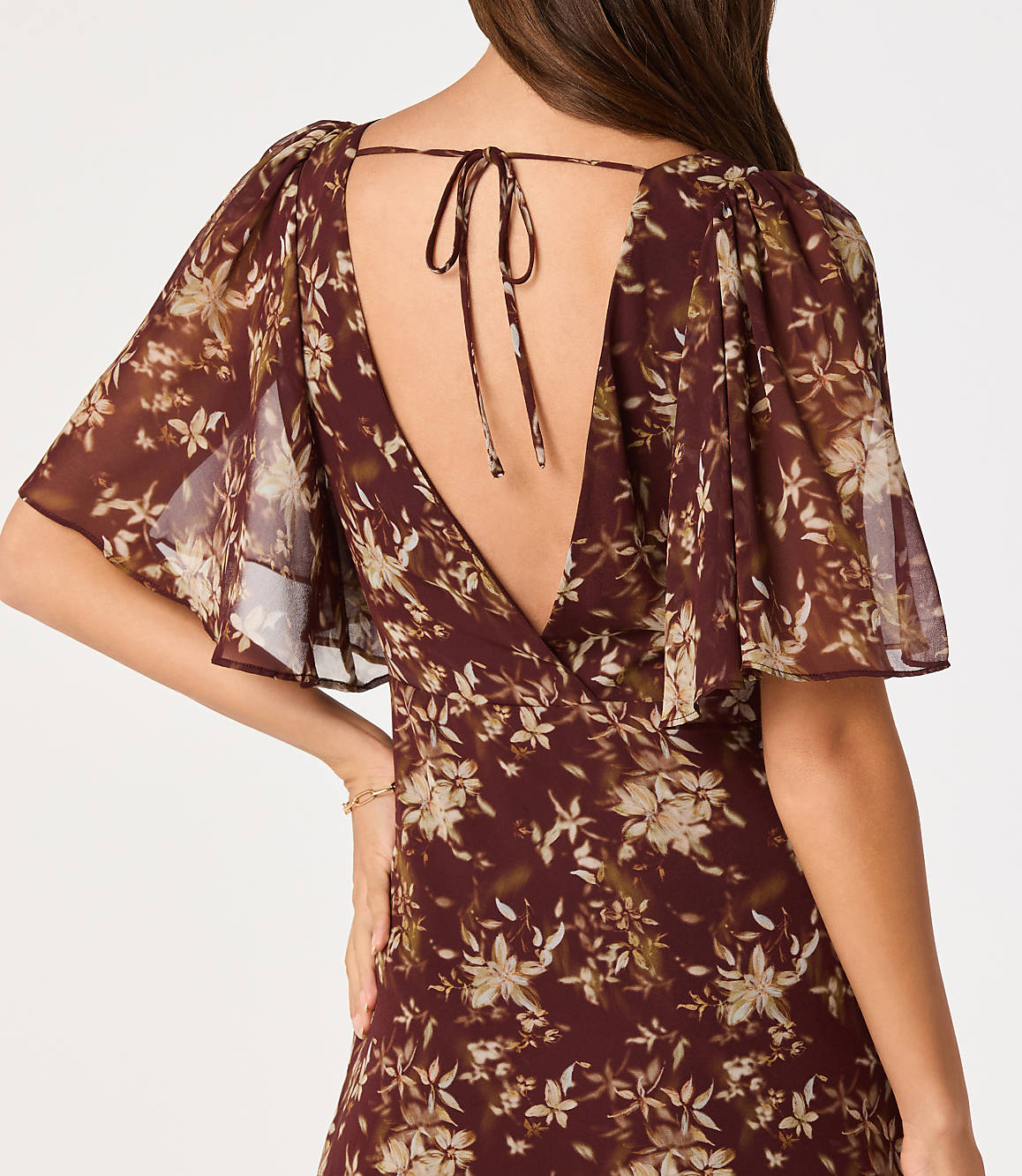 A woman in a floral print dress with flutter sleeves and a tie-back neckline stands against a plain background.
