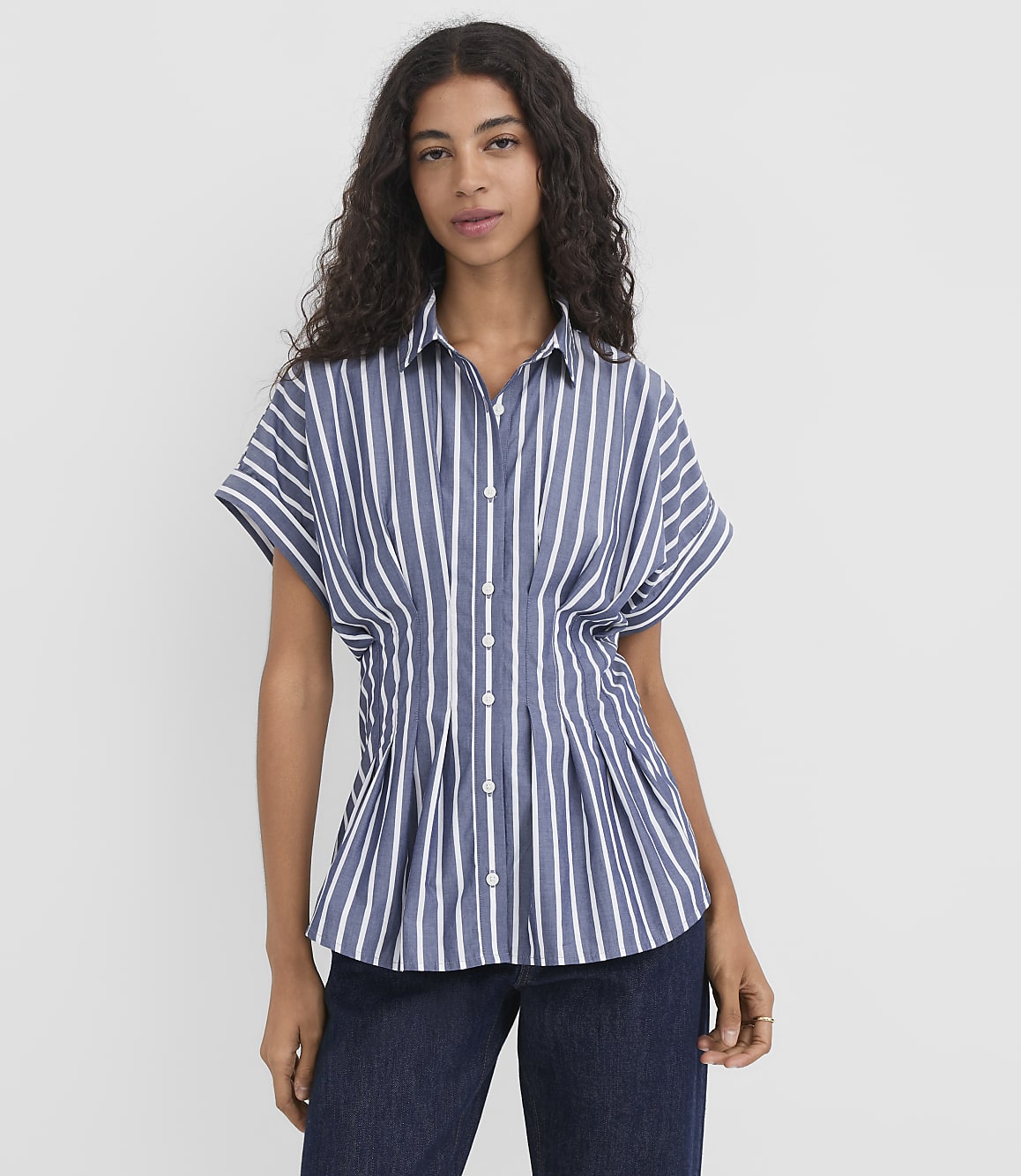A woman with dark, curly hair stands in the foreground wearing a blue and white striped shirt and dark jeans, against a plain white background.