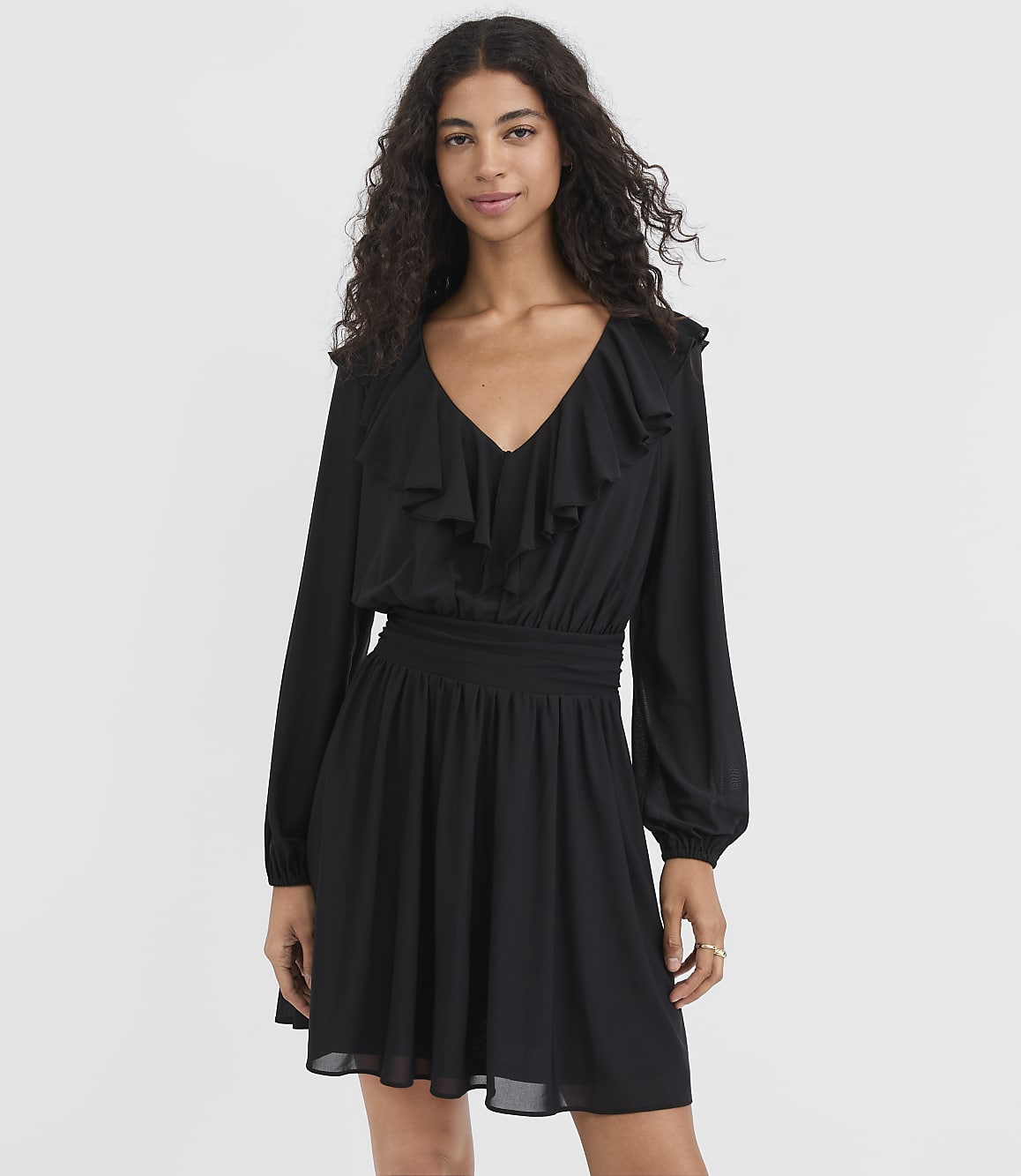 A woman in a black ruffled dress stands against a plain white background.
