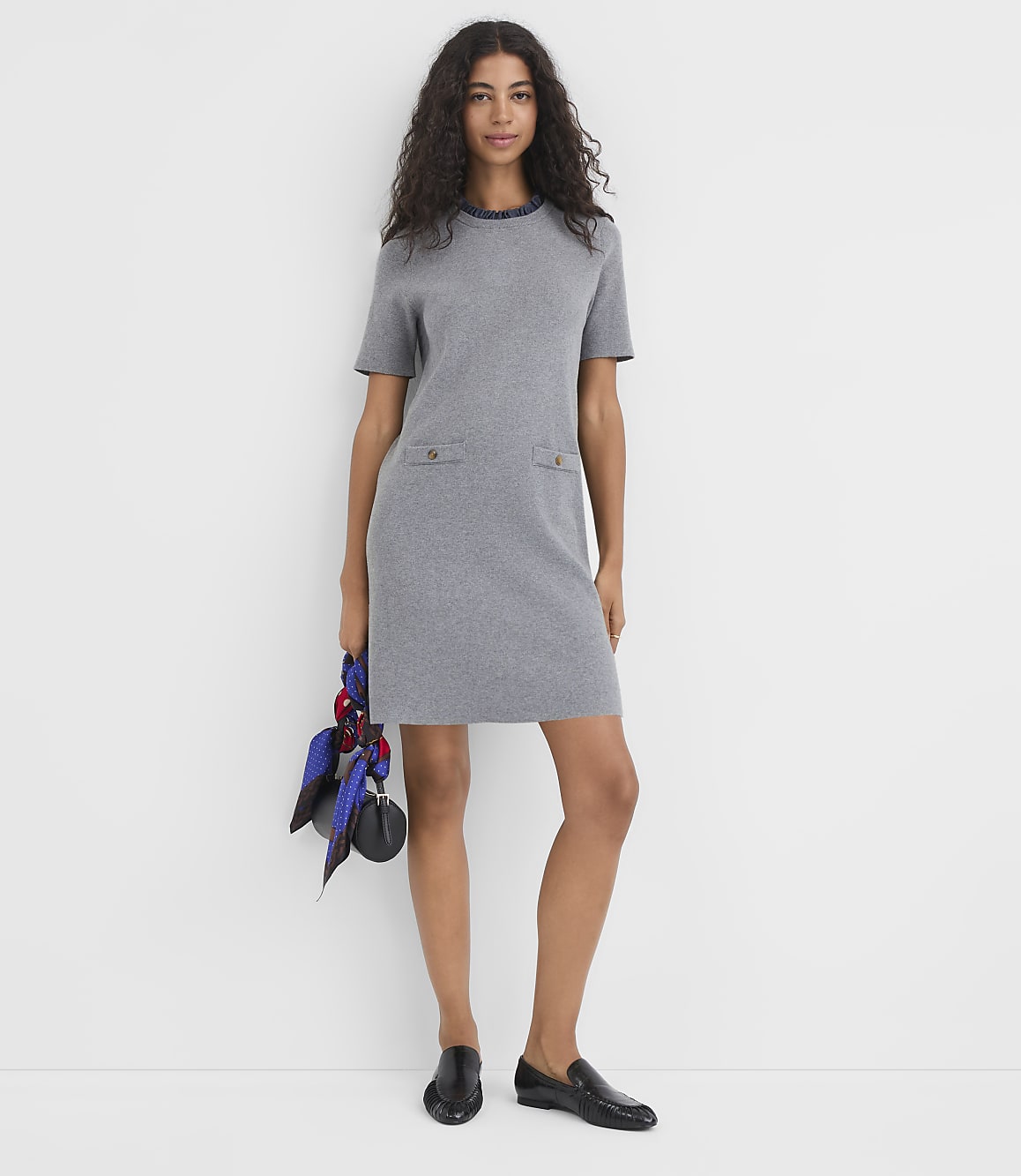 A woman in a grey dress and black loafers stands against a plain white background, holding a patterned scarf and a small black purse.