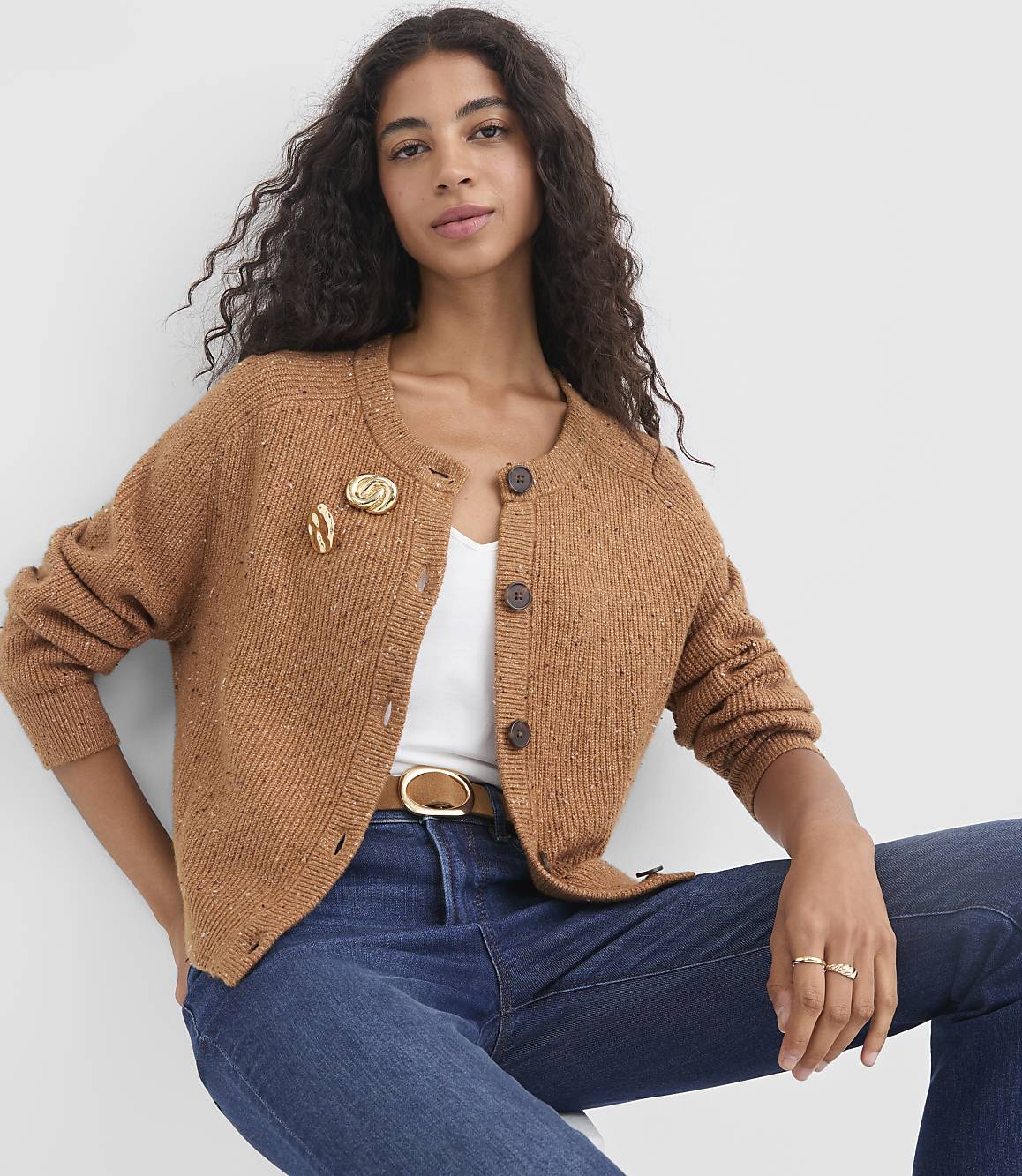 A woman in a brown cardigan and blue jeans sits against a plain white background.
