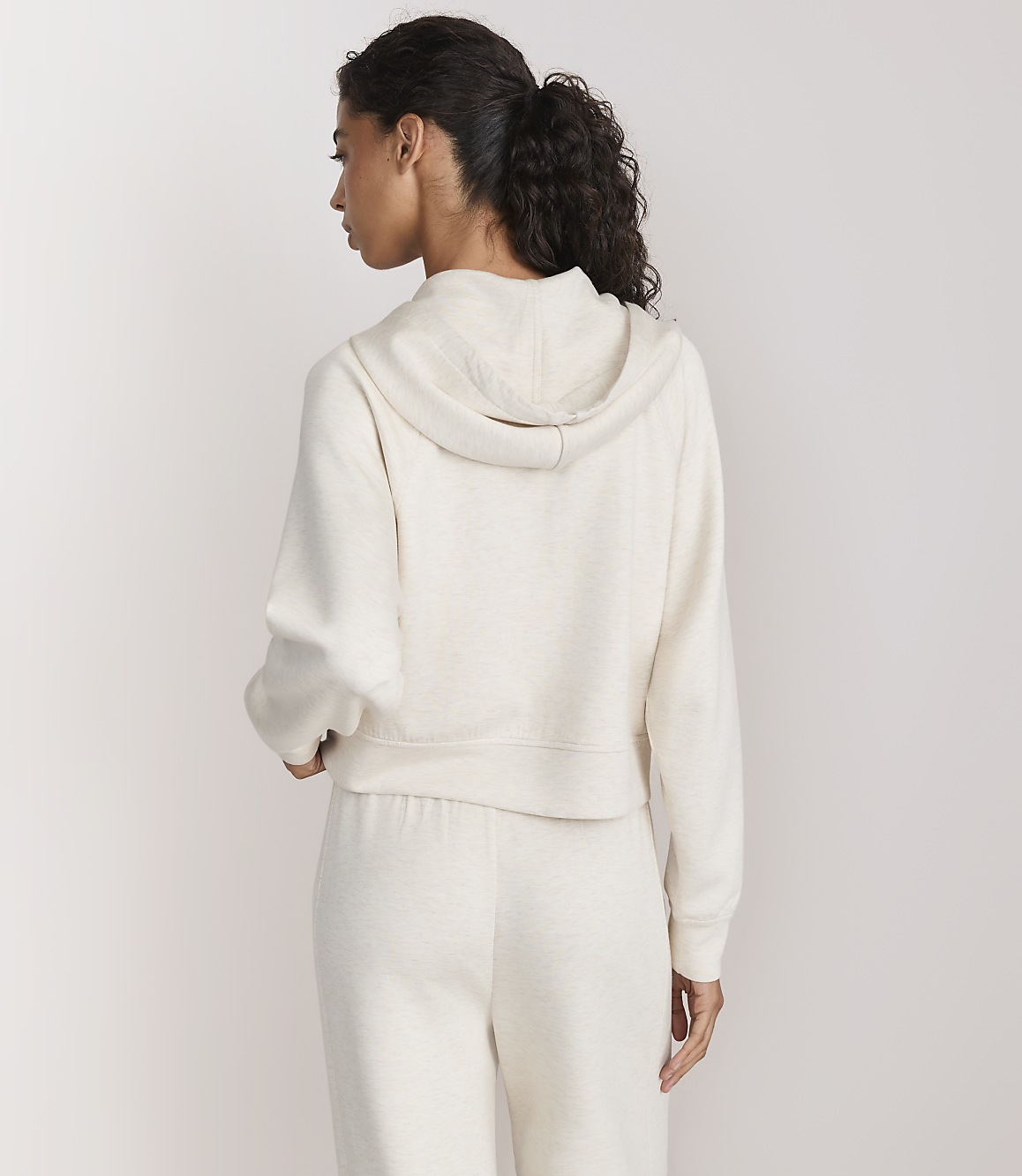A woman in a cream-colored hoodie and sweatpants stands against a plain white background.