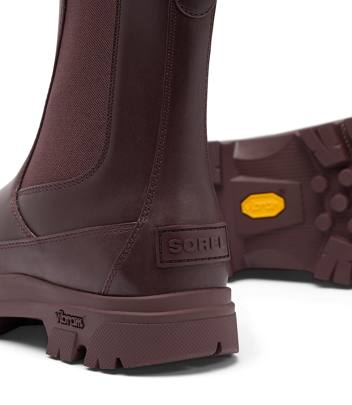 A close-up of burgundy leather Chelsea boots with thick soles and a Vibram logo is set against a white background.
