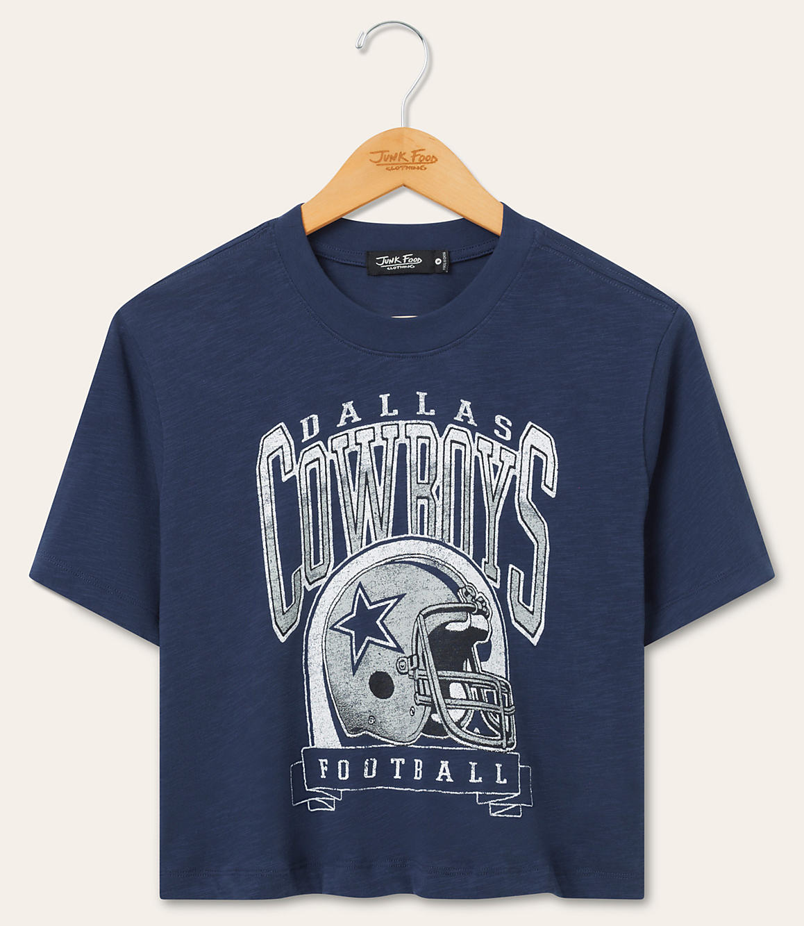 A navy blue Dallas Cowboys t-shirt with a graphic of a helmet and team name hangs on a wooden hanger against a plain beige background.