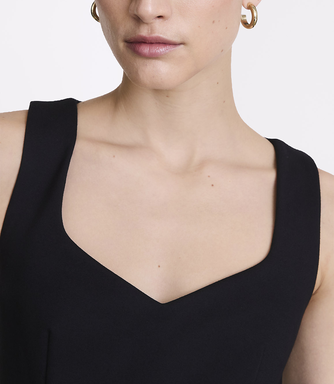 A woman with gold hoop earrings wears a black dress with a sweetheart neckline, against a plain white background.
