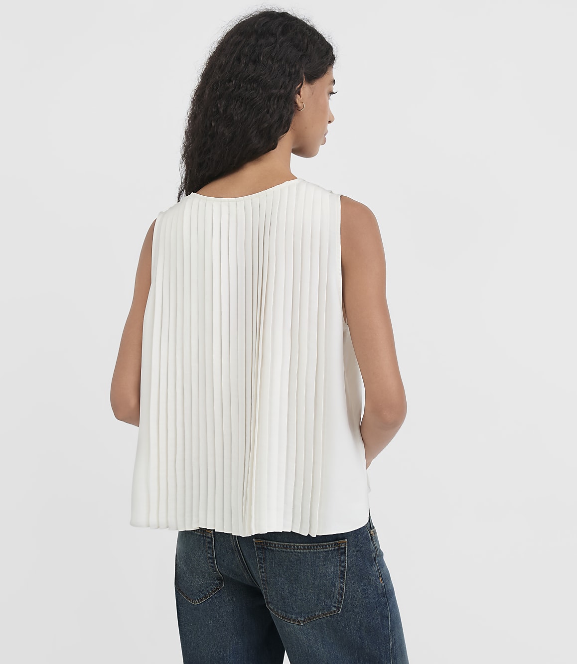 A woman in a white pleated top and blue jeans stands against a plain white background.