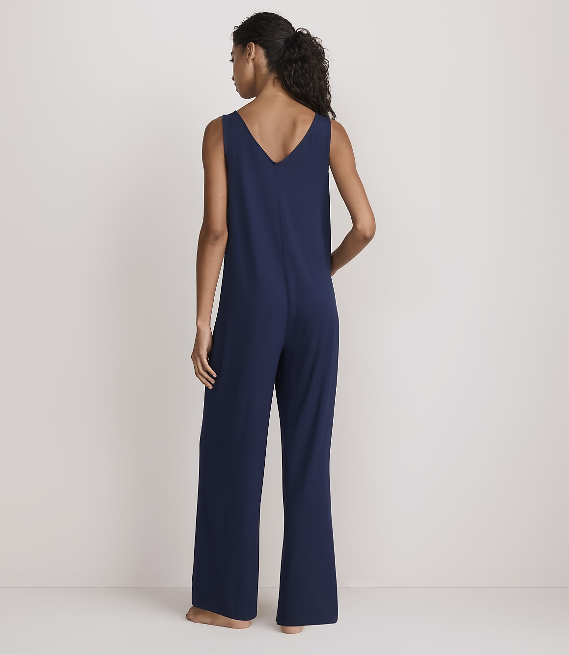 A woman in a navy blue jumpsuit stands with her back to the viewer against a plain white background.