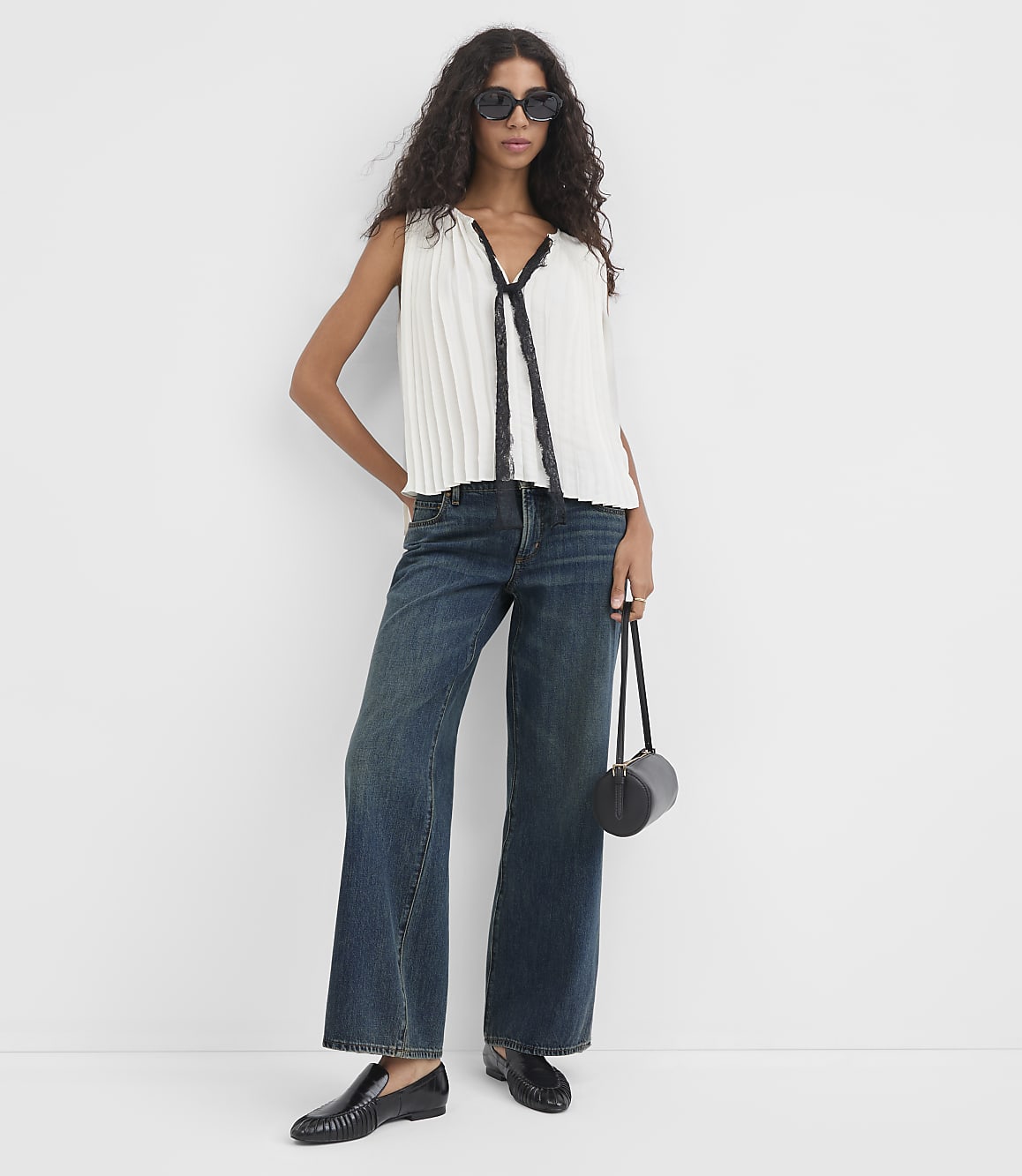 A woman in a white pleated top and dark jeans stands against a plain white background, holding a black cylindrical bag.