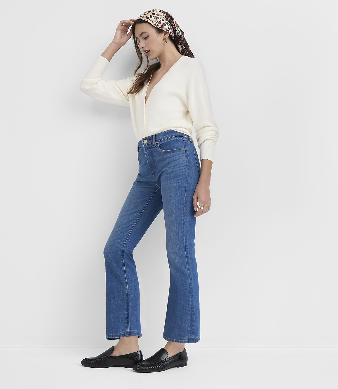A woman in a cream cardigan and blue jeans stands against a plain white background.