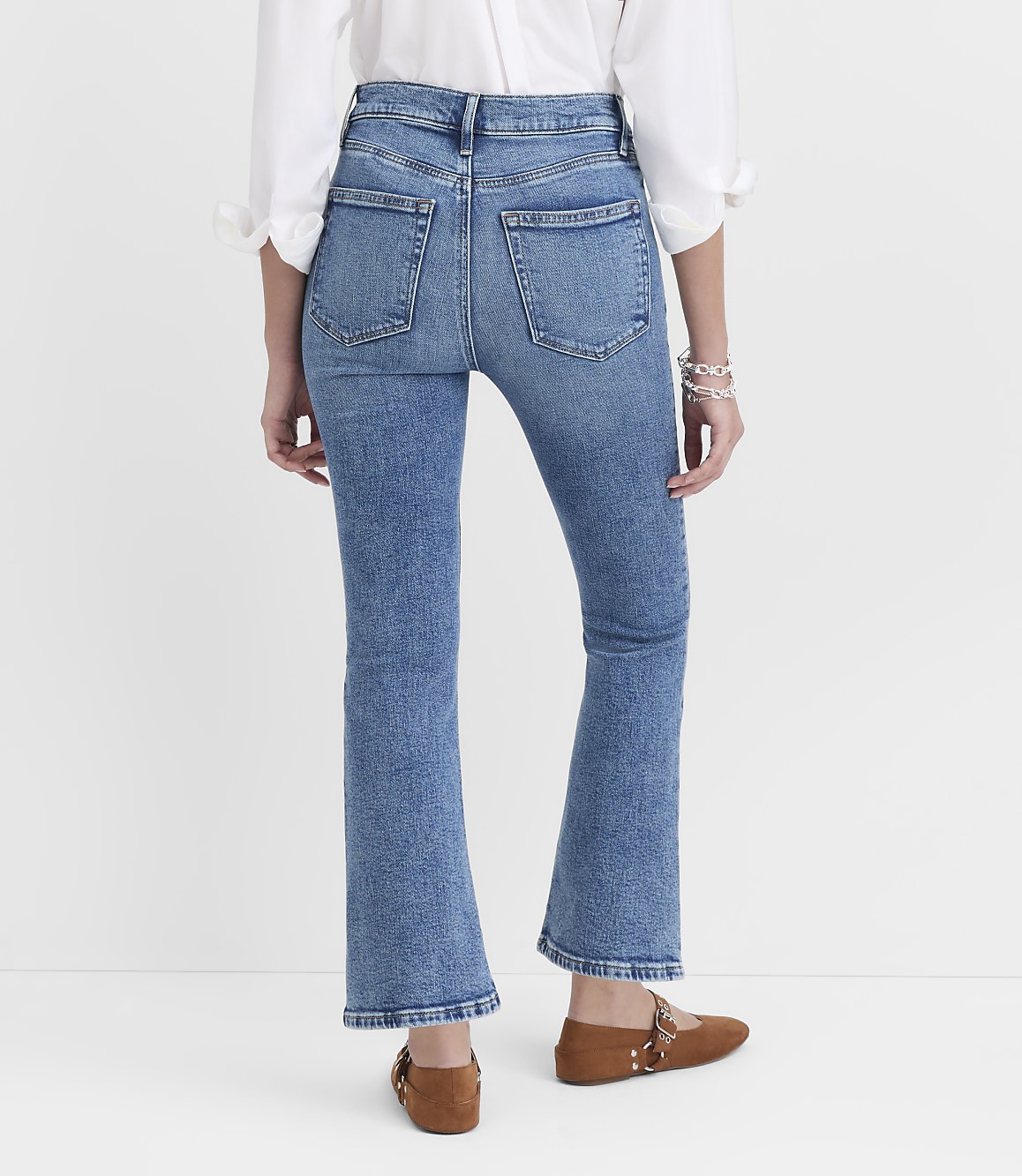 A person wearing flared blue jeans and brown flats stands against a plain white background.