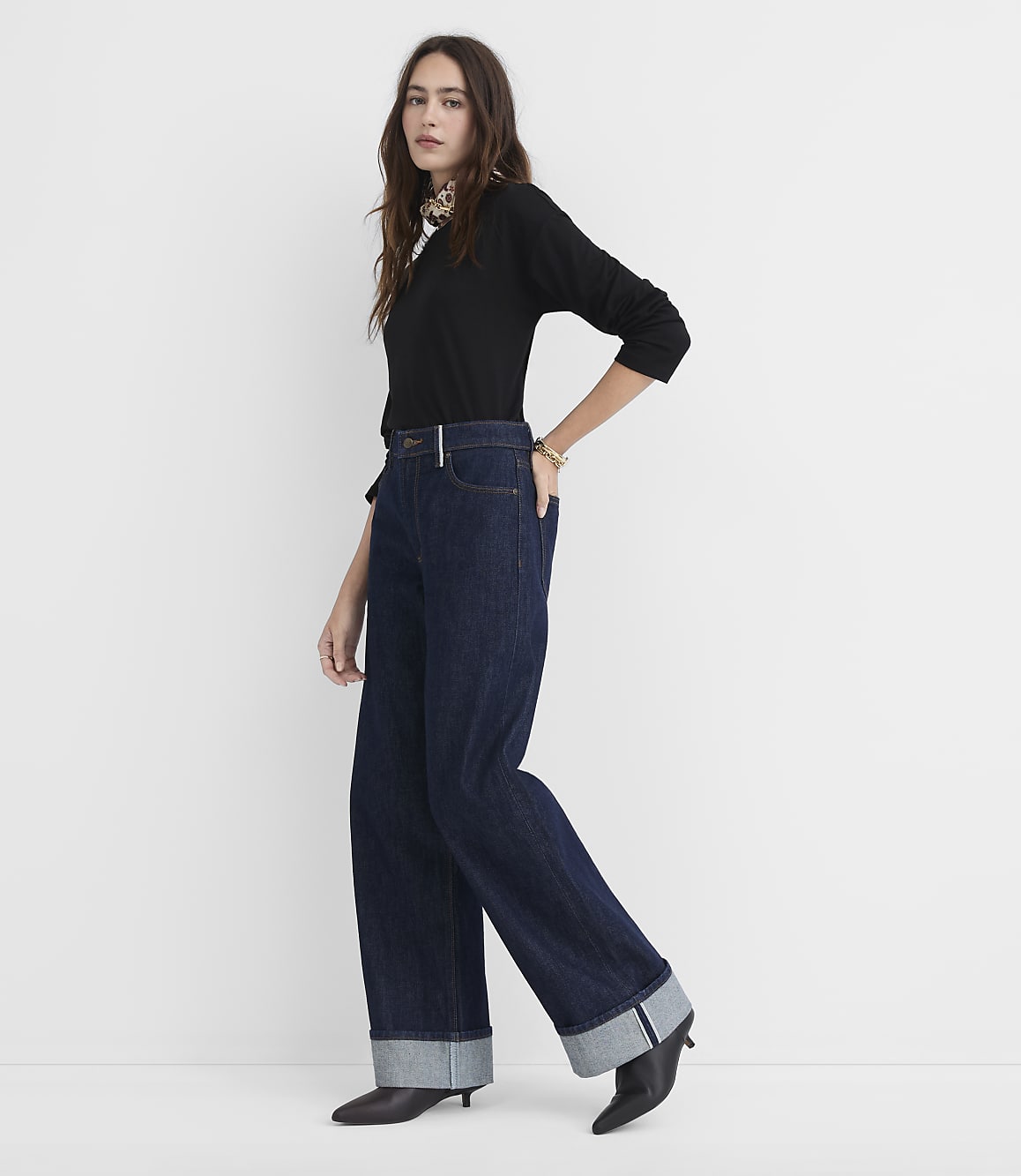 A woman in a black top and wide-leg jeans stands against a plain white background.