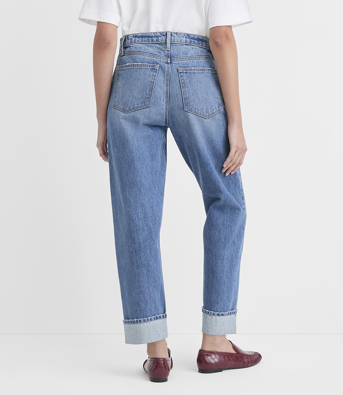 A person wearing cuffed denim jeans and burgundy loafers stands against a plain white background.