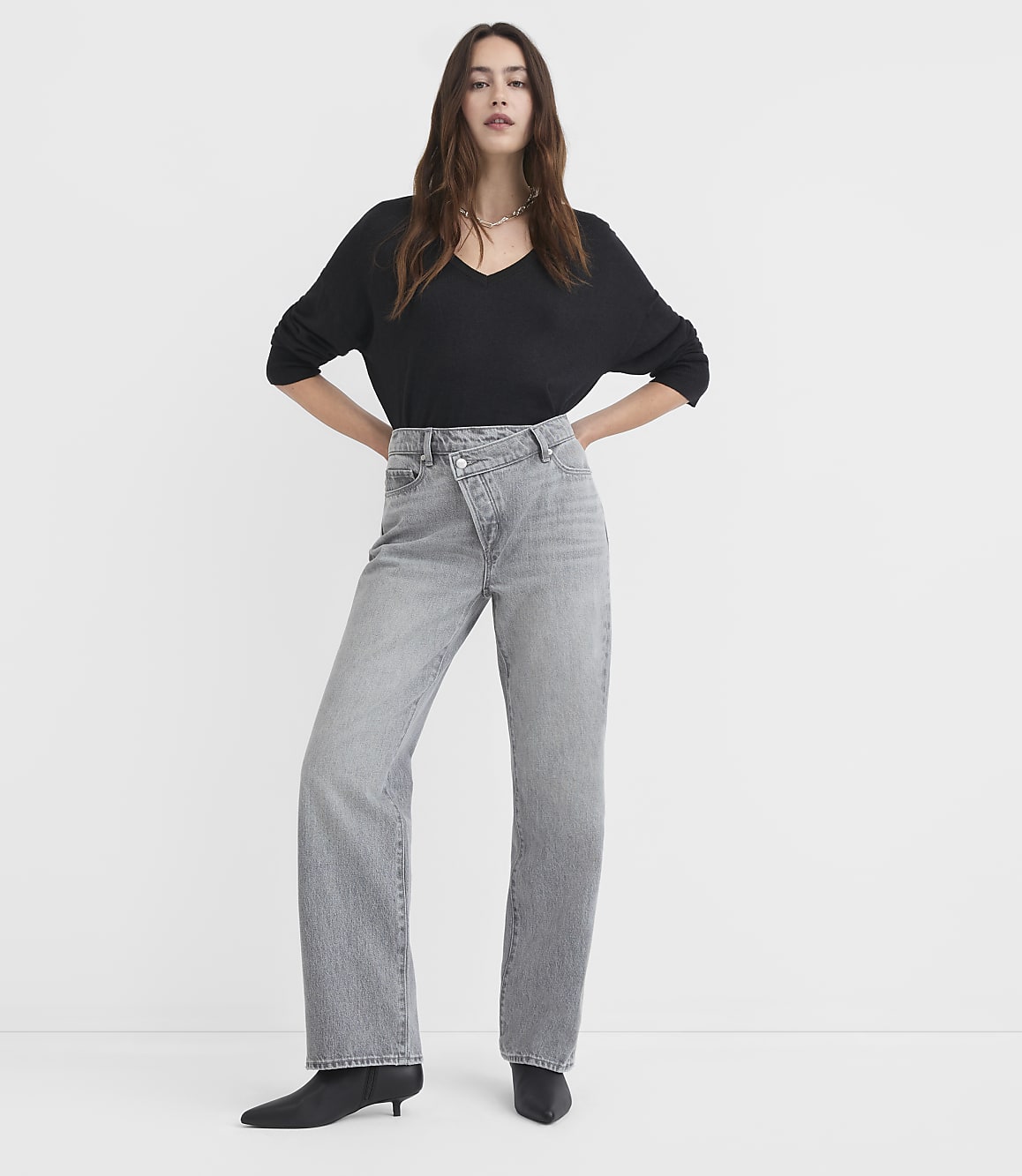 A woman in a black v-neck shirt and grey criss-cross jeans stands against a plain white background.