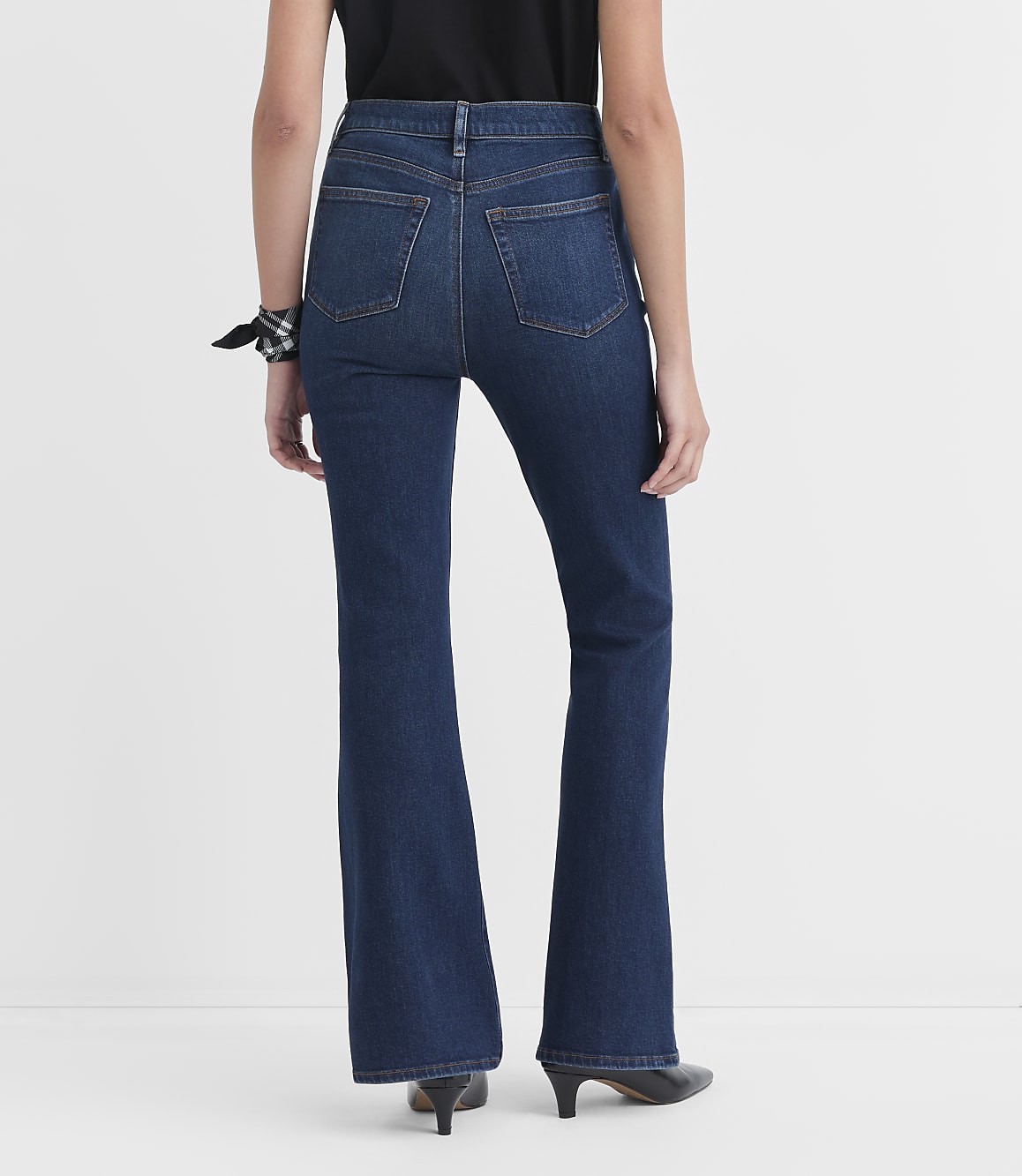 A person wearing dark wash flare jeans and black heels stands against a plain white background.