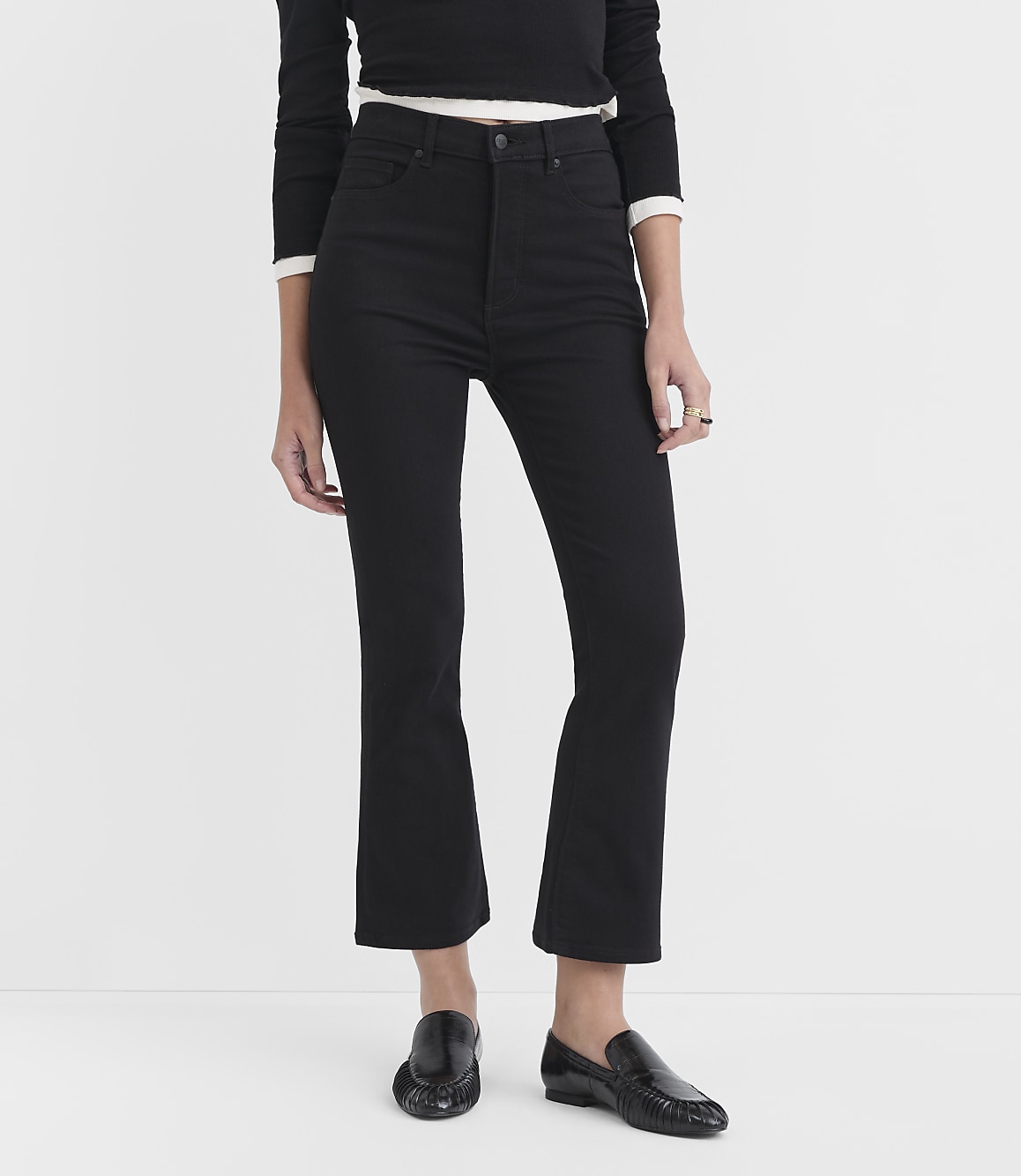 A person wearing black flared jeans and black loafers stands against a plain white background.