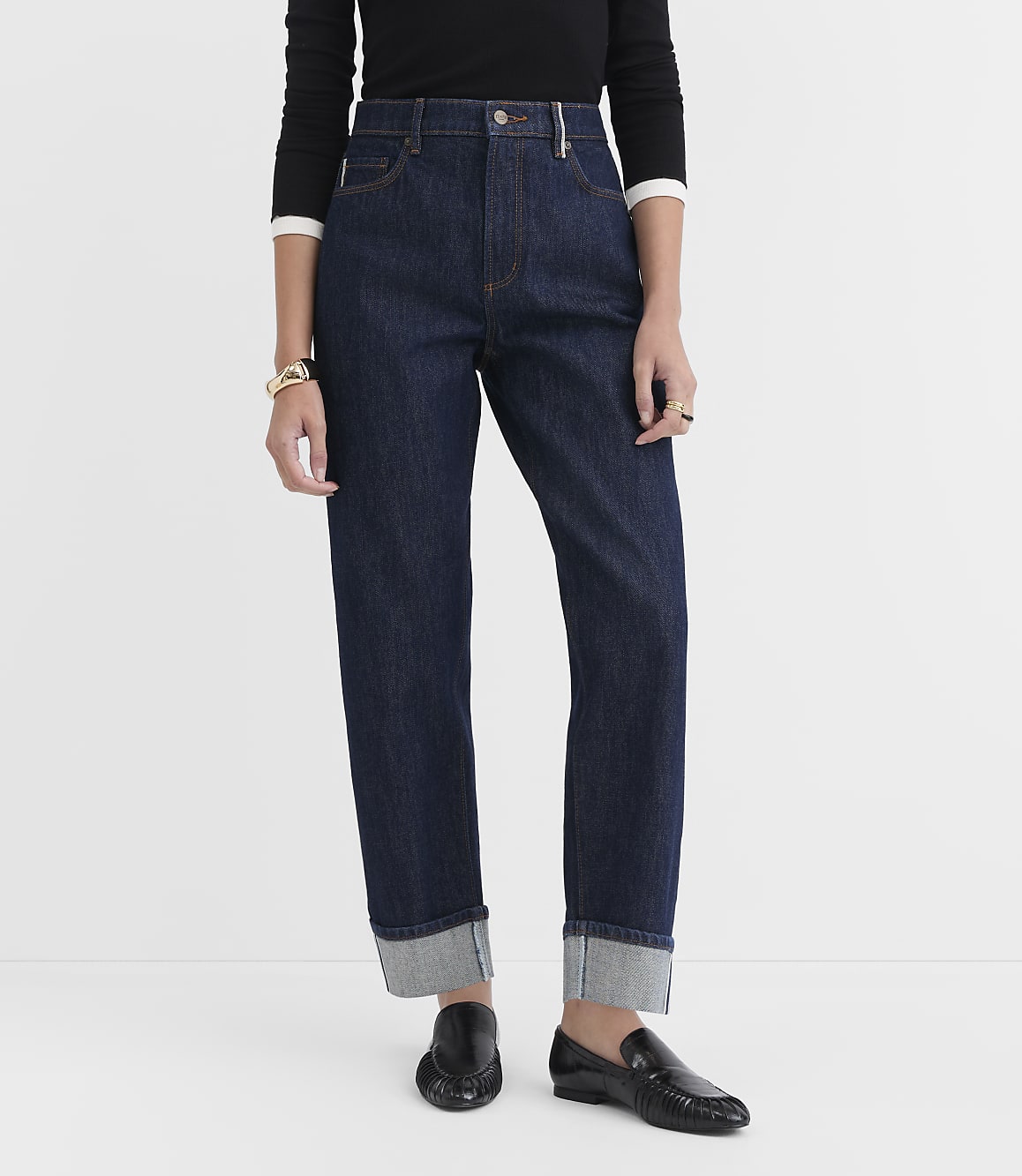 A person stands in dark wash cuffed jeans and black loafers against a plain white background.