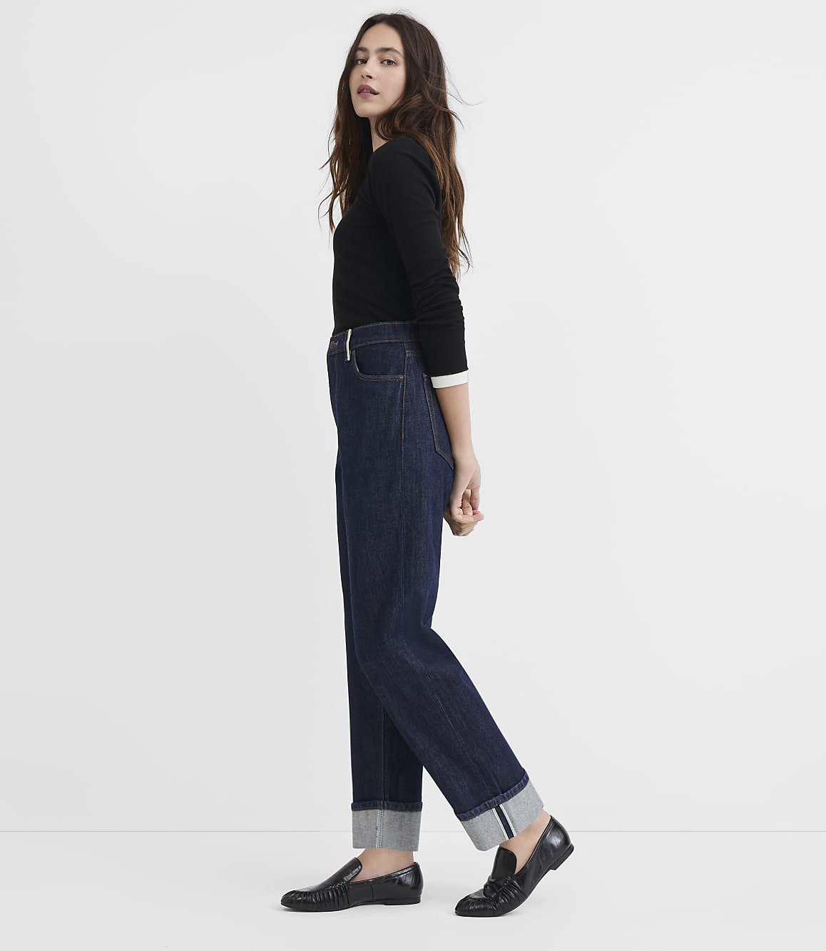 A woman in a black sweater and dark wash cuffed jeans stands against a plain white background.