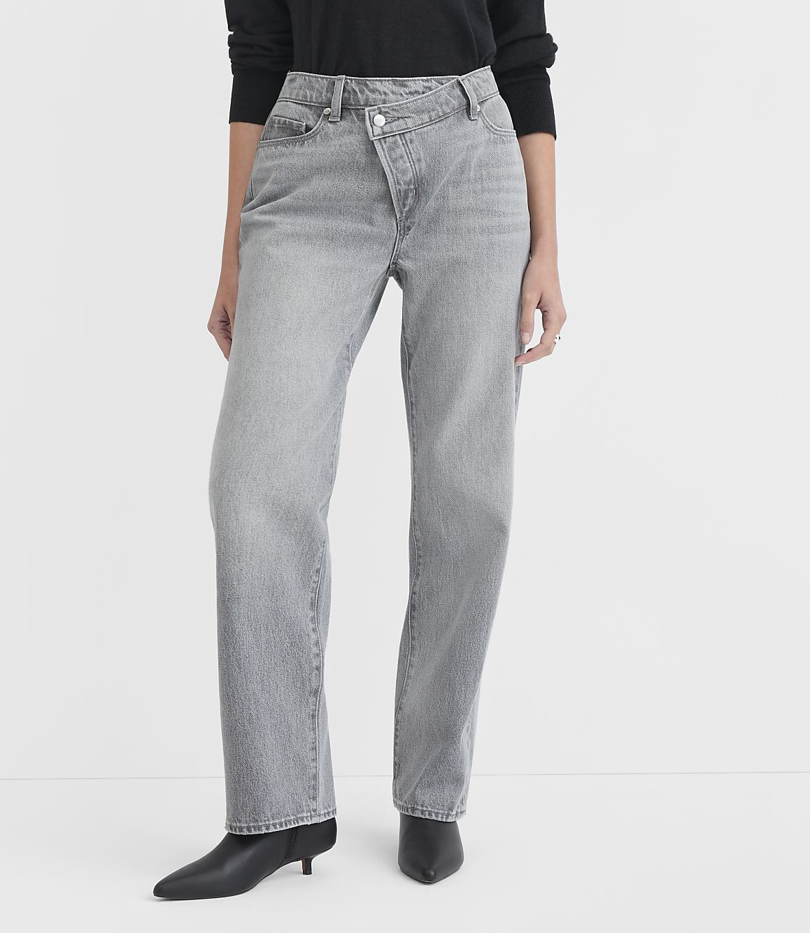 A person models light gray, asymmetrical-waist jeans and black pointed-toe boots against a plain white background.