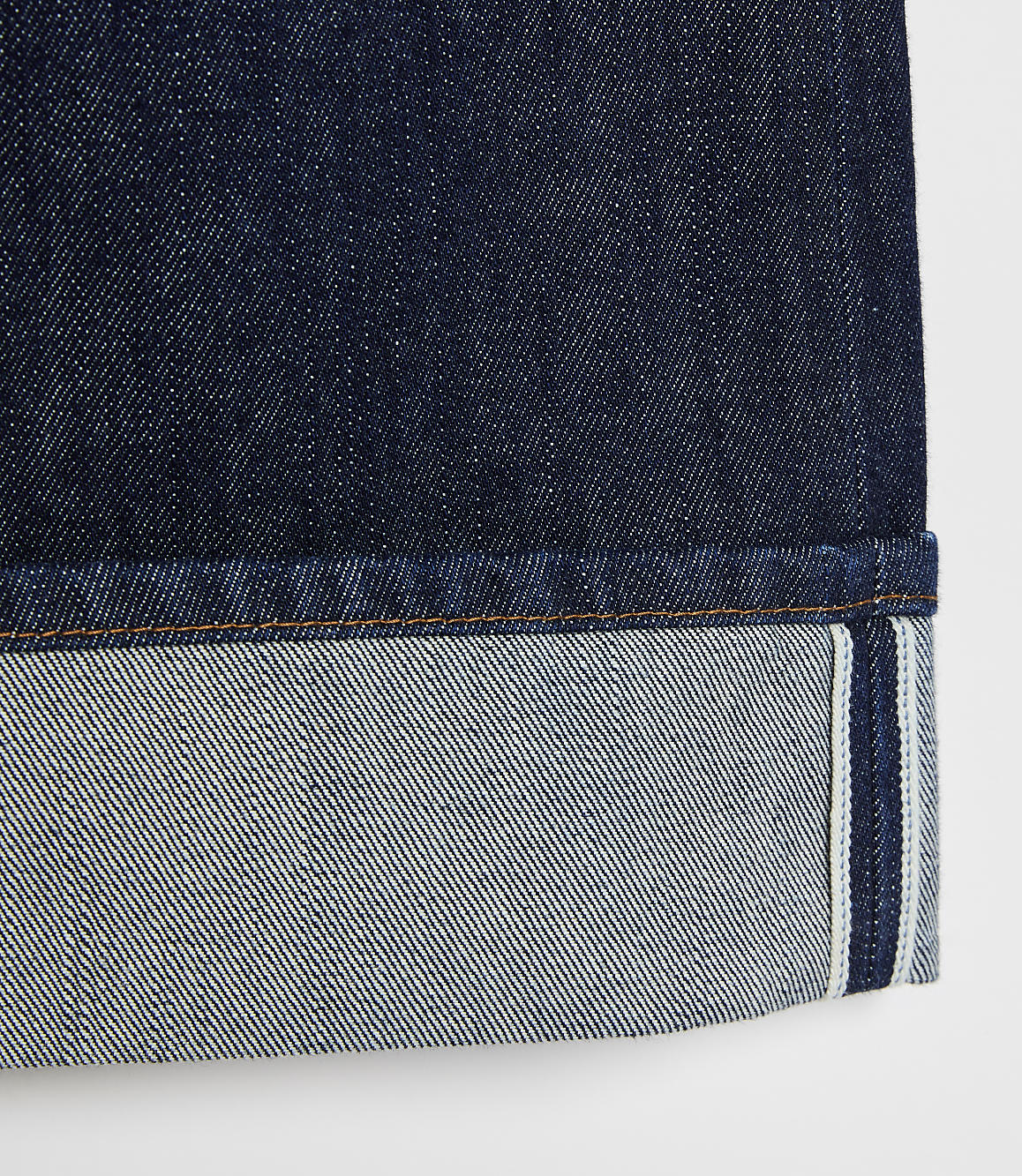 A close-up of dark indigo denim jeans with a cuffed hem showing the lighter interior fabric and selvedge edge against a plain white background.
