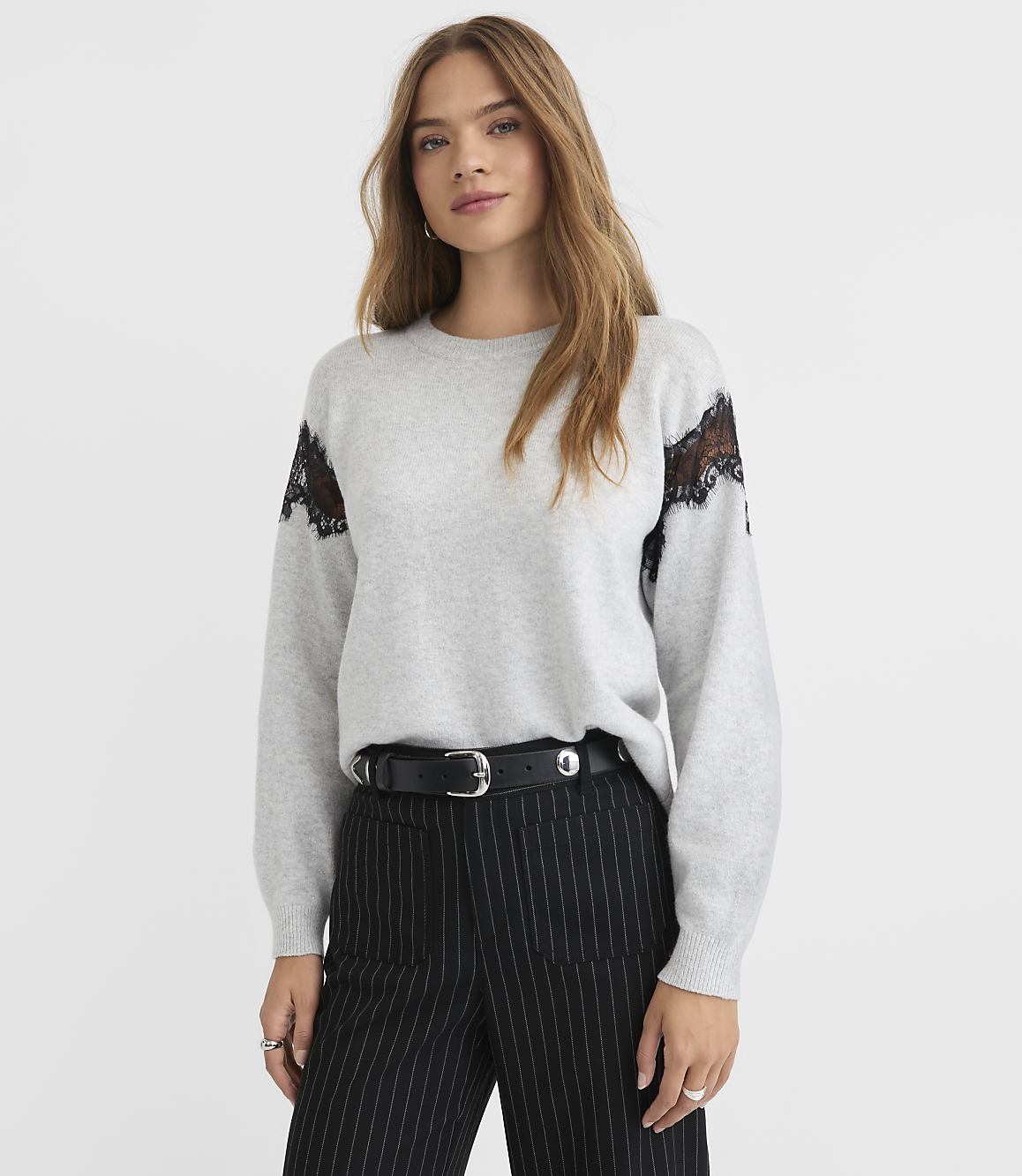 A woman in a grey sweater with black lace accents on the shoulders and pinstripe pants stands against a plain grey background.