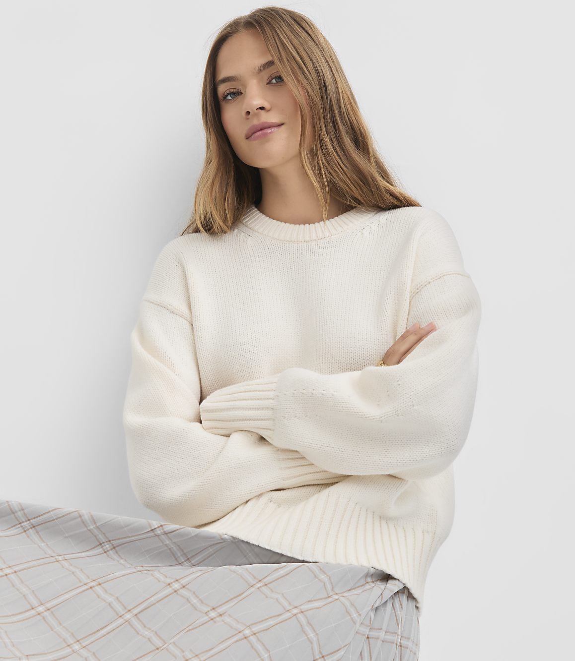 A woman in a cream sweater and plaid skirt poses with her arms crossed against a plain white background.