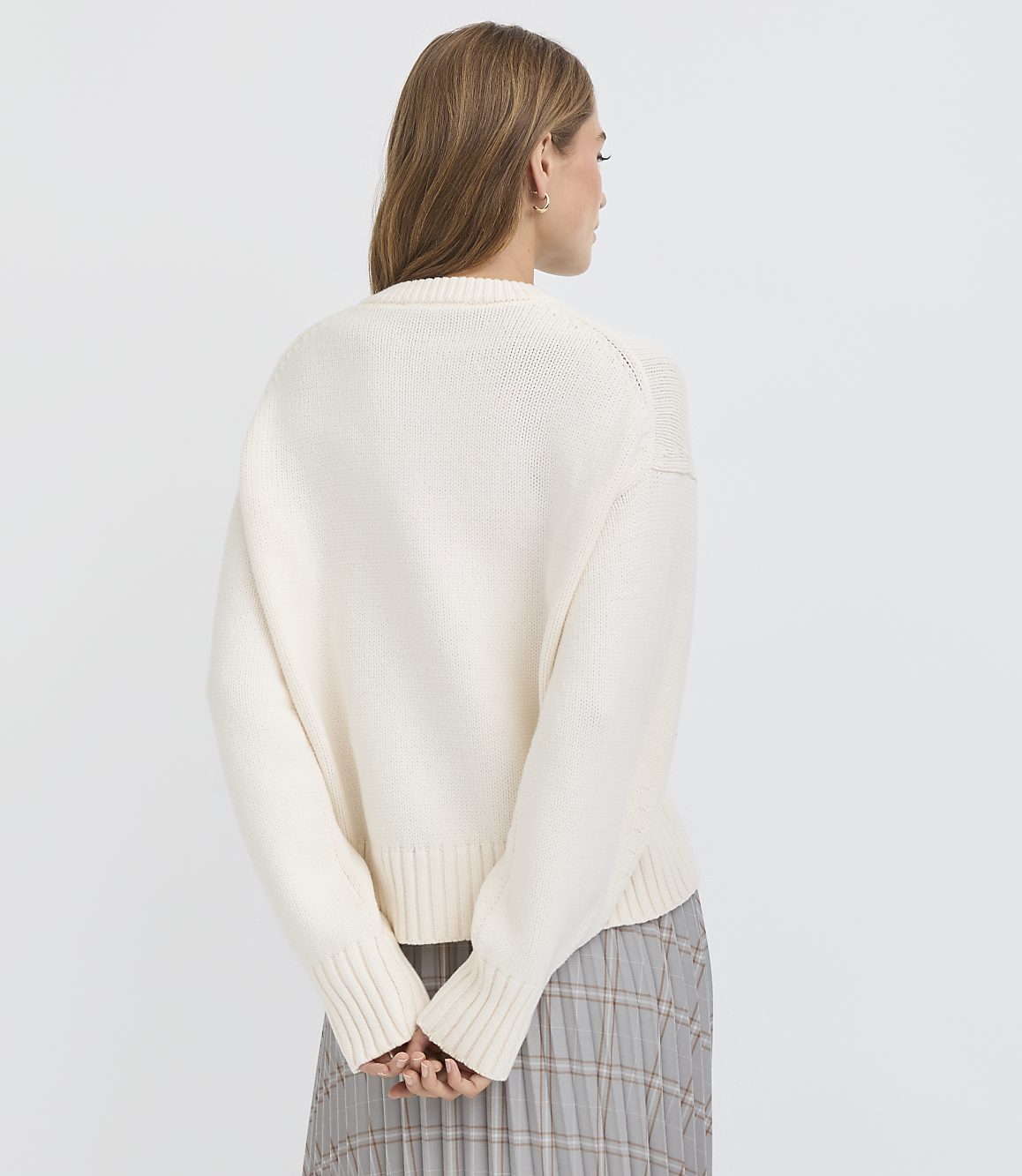 A woman in a cream-colored sweater and plaid skirt stands with her back to the camera against a plain white background.