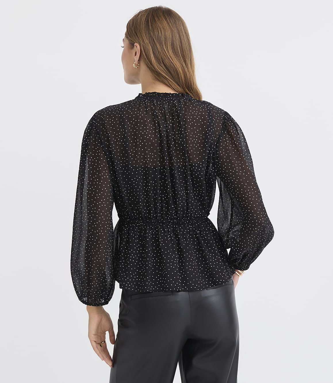 A woman in a black polka dot blouse and leather pants stands against a plain white background.