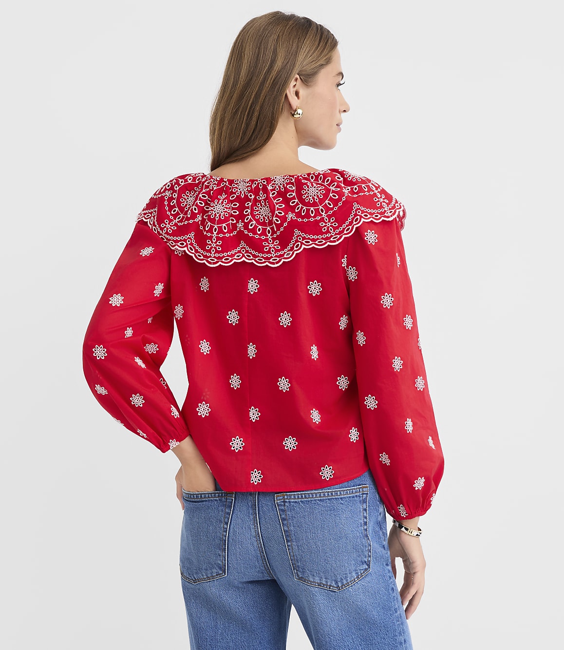 A woman in a red embroidered blouse and blue jeans stands against a plain white background.