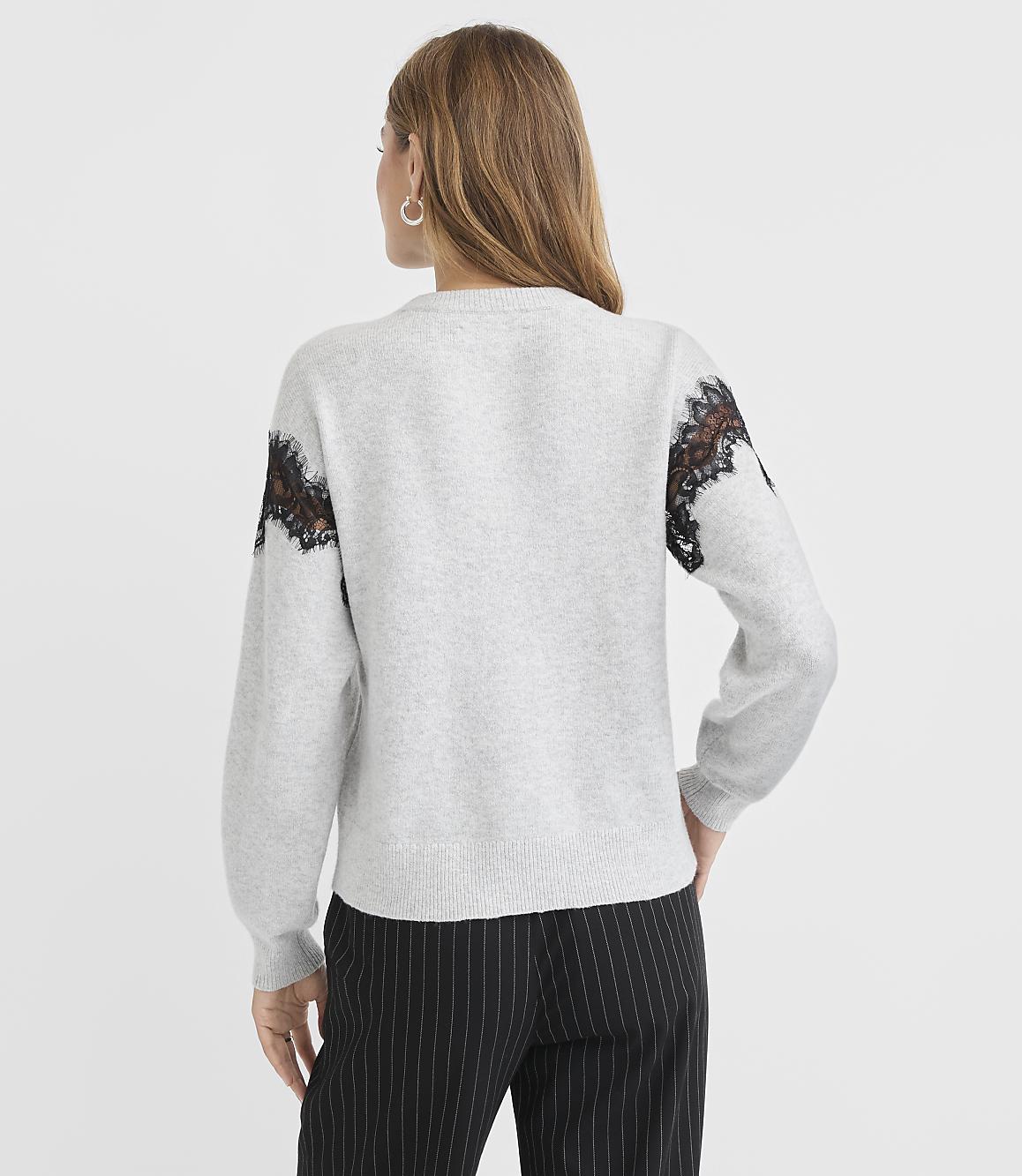 A woman in a light gray sweater with black lace accents on the shoulders and pinstripe pants stands against a plain gray background.