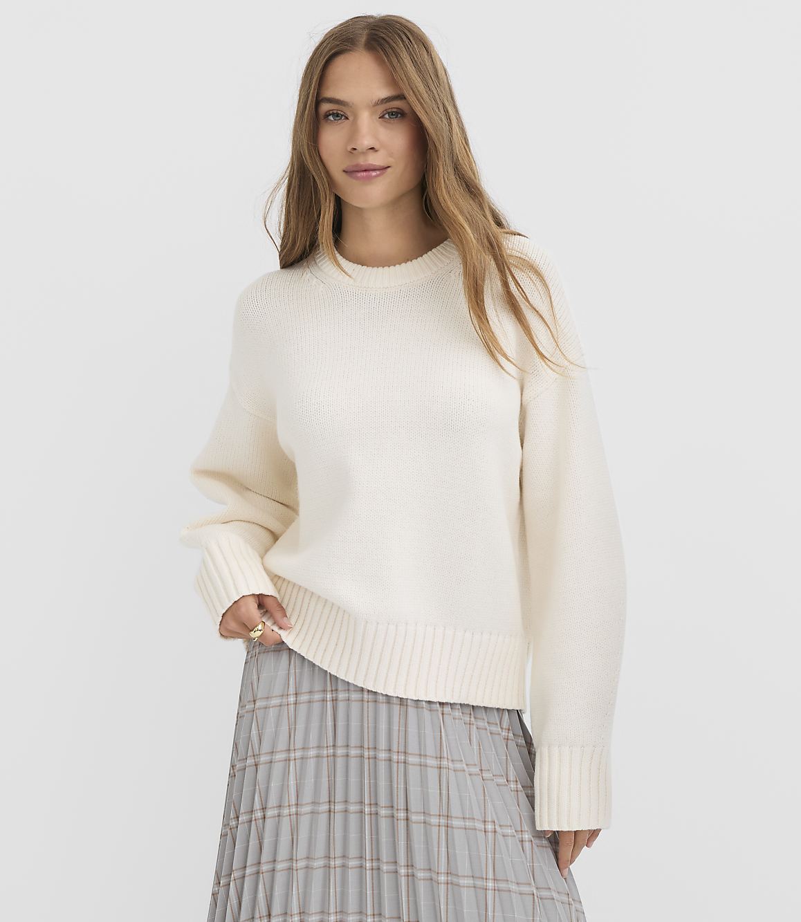 A young woman in a cream sweater and plaid skirt stands against a plain white background.