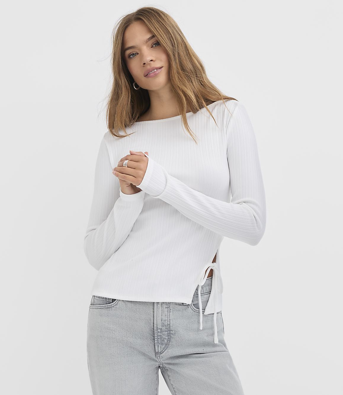 A young woman in a white ribbed top and light wash jeans stands against a plain white background.