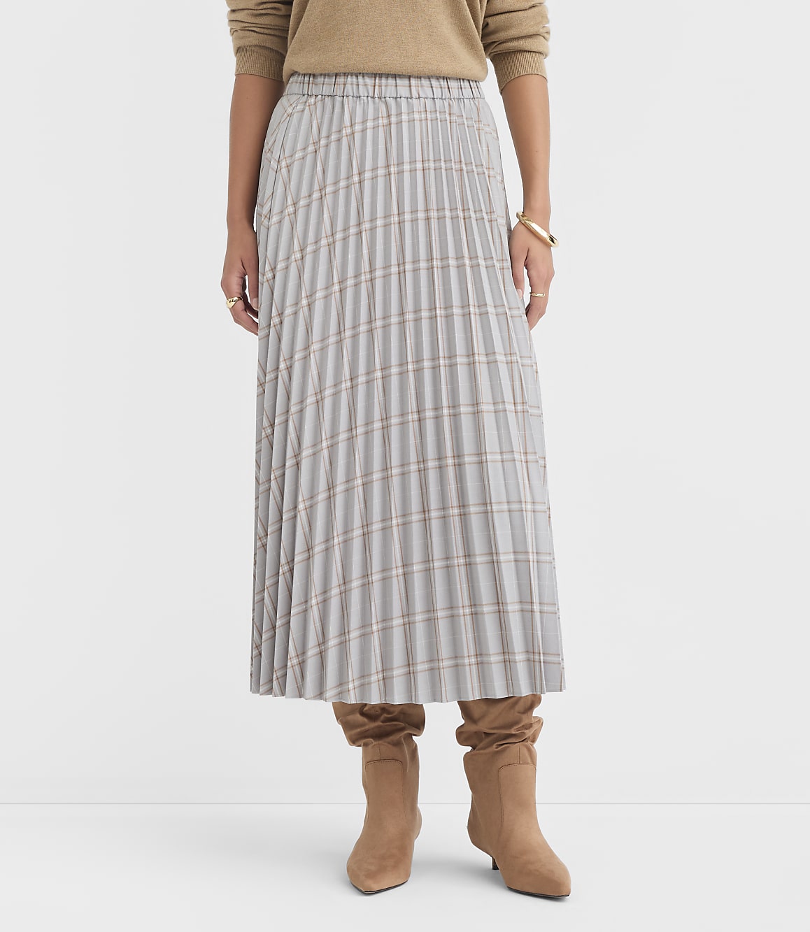 A woman in a plaid pleated skirt and brown boots stands against a plain white background.