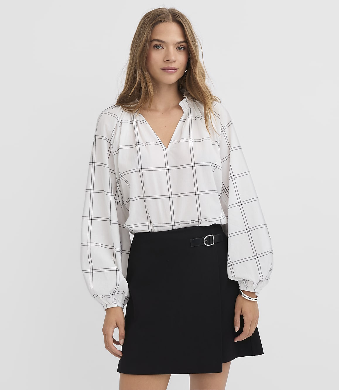 A woman in a white plaid blouse and black skirt stands against a plain white background.