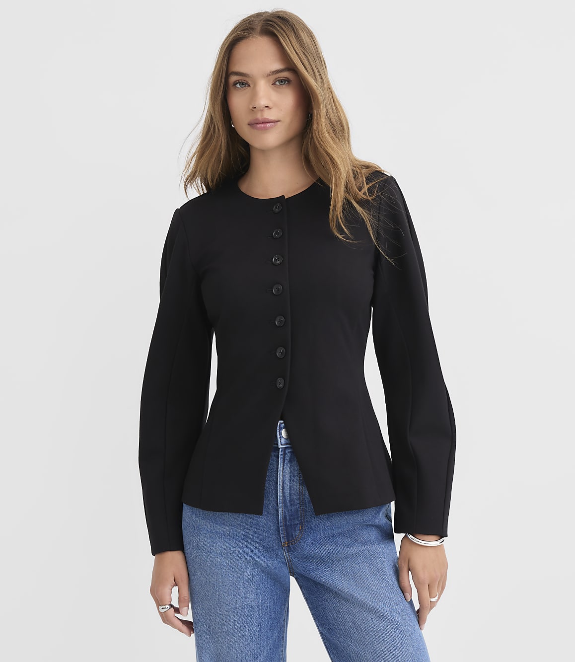A woman in a black jacket and blue jeans stands against a plain white background.