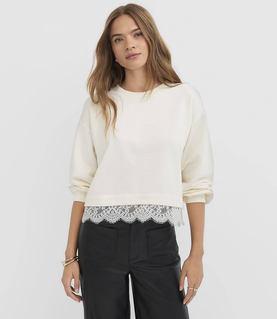 A young woman in a cream sweatshirt with a lace hem stands against a plain white background.
