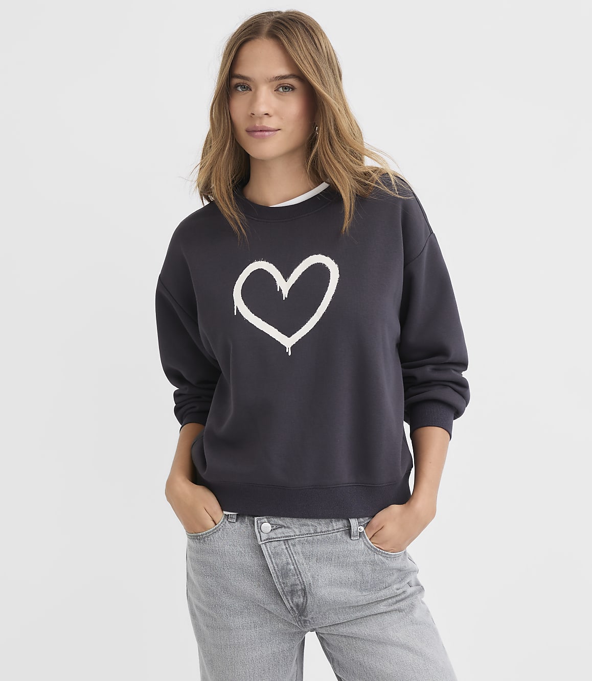 A young woman in a dark sweatshirt with a white heart graphic stands against a plain white background.