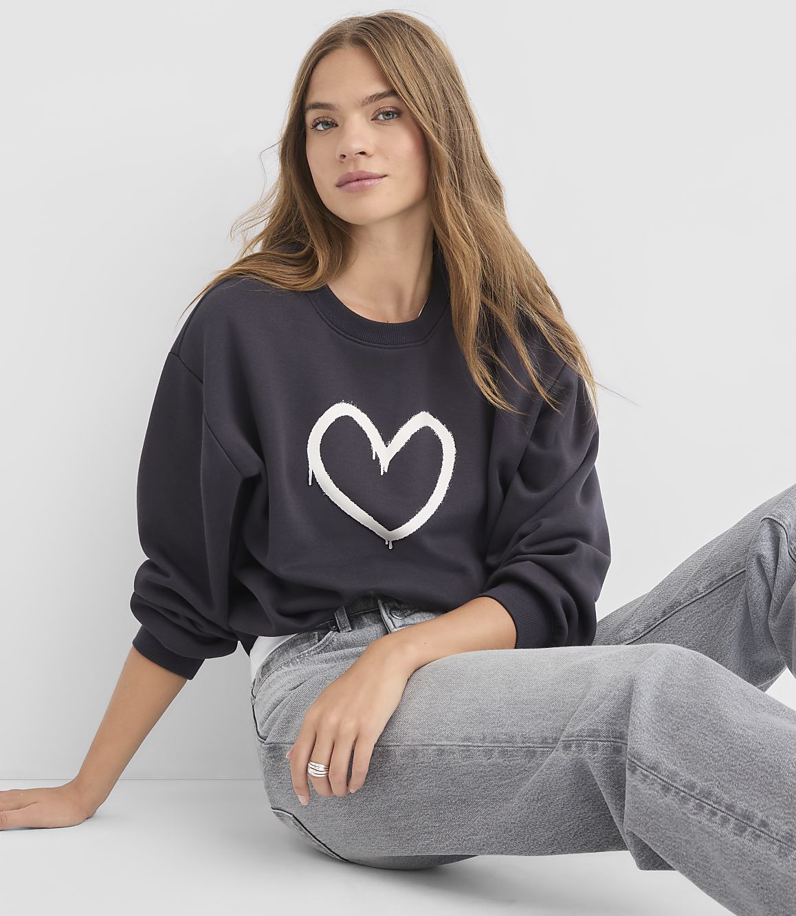 A young woman in a dark sweatshirt with a white heart graphic and grey jeans sits against a plain white background.