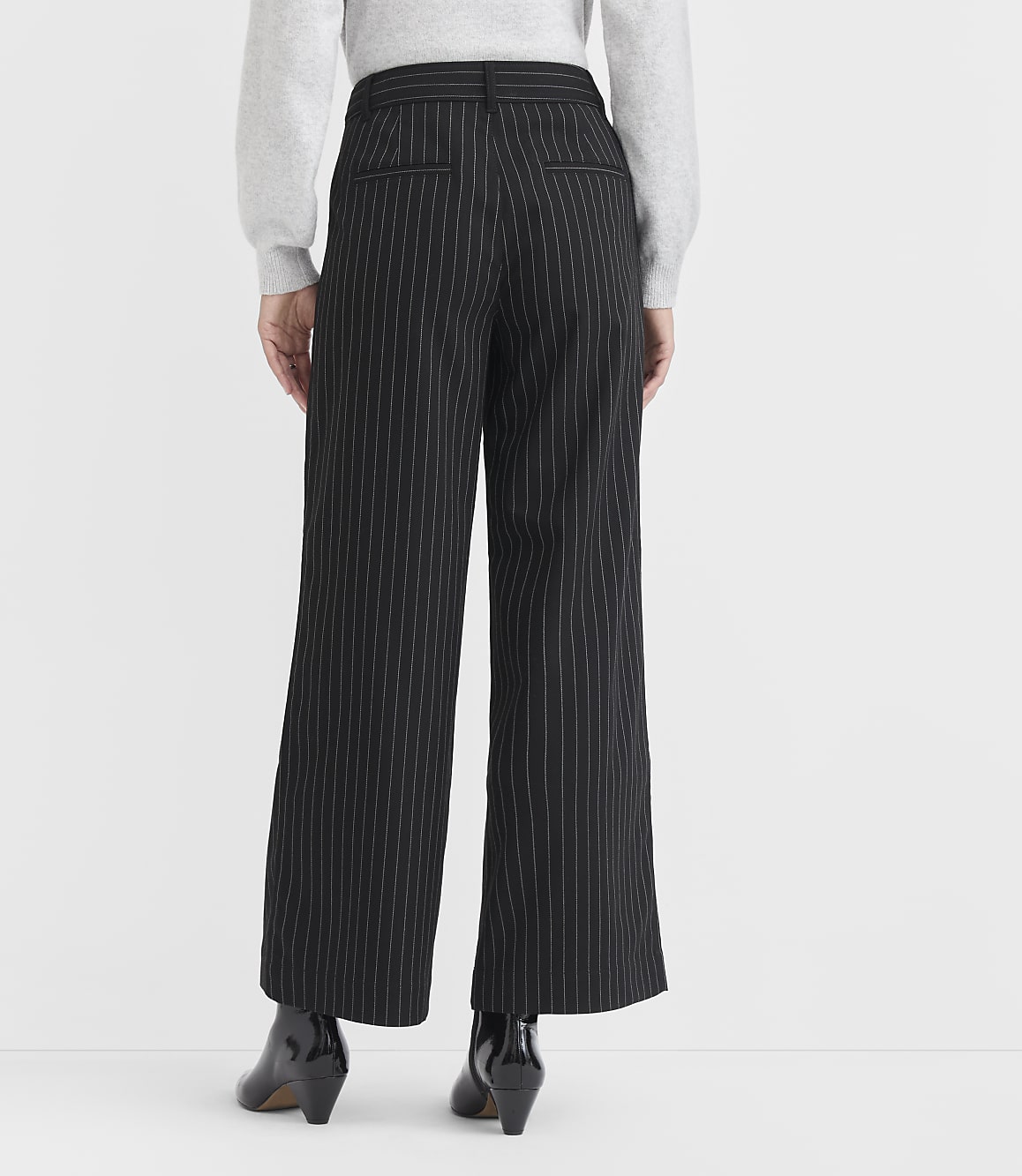 A person wearing black pinstripe wide-leg pants and black heeled boots stands against a plain white background.