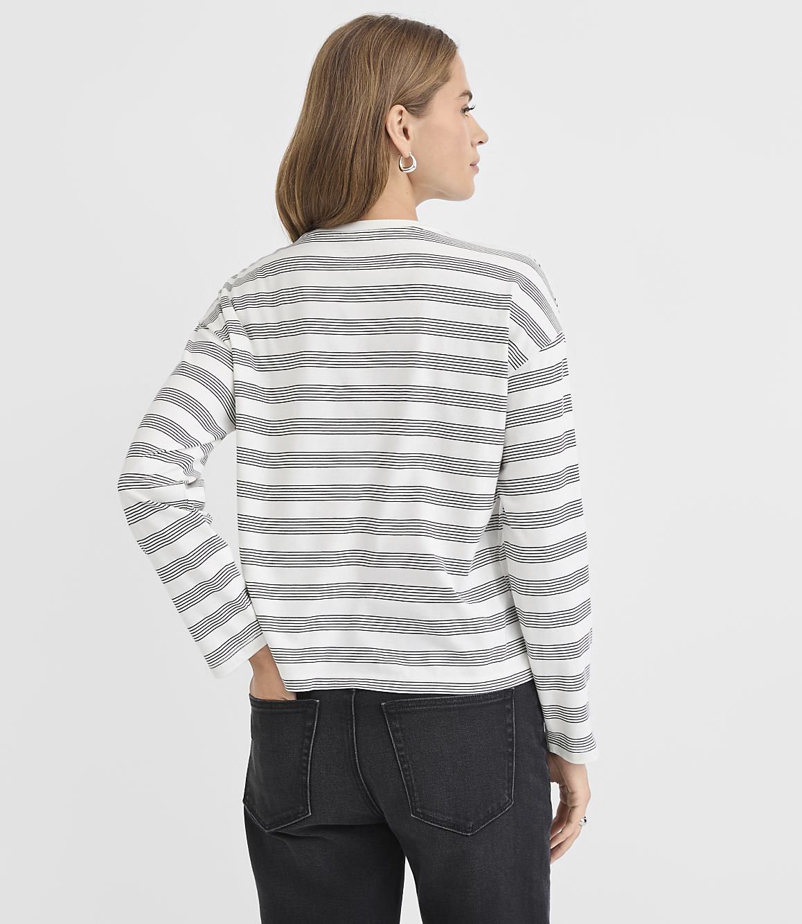 A woman in a striped long-sleeved shirt and dark jeans stands against a plain white background.