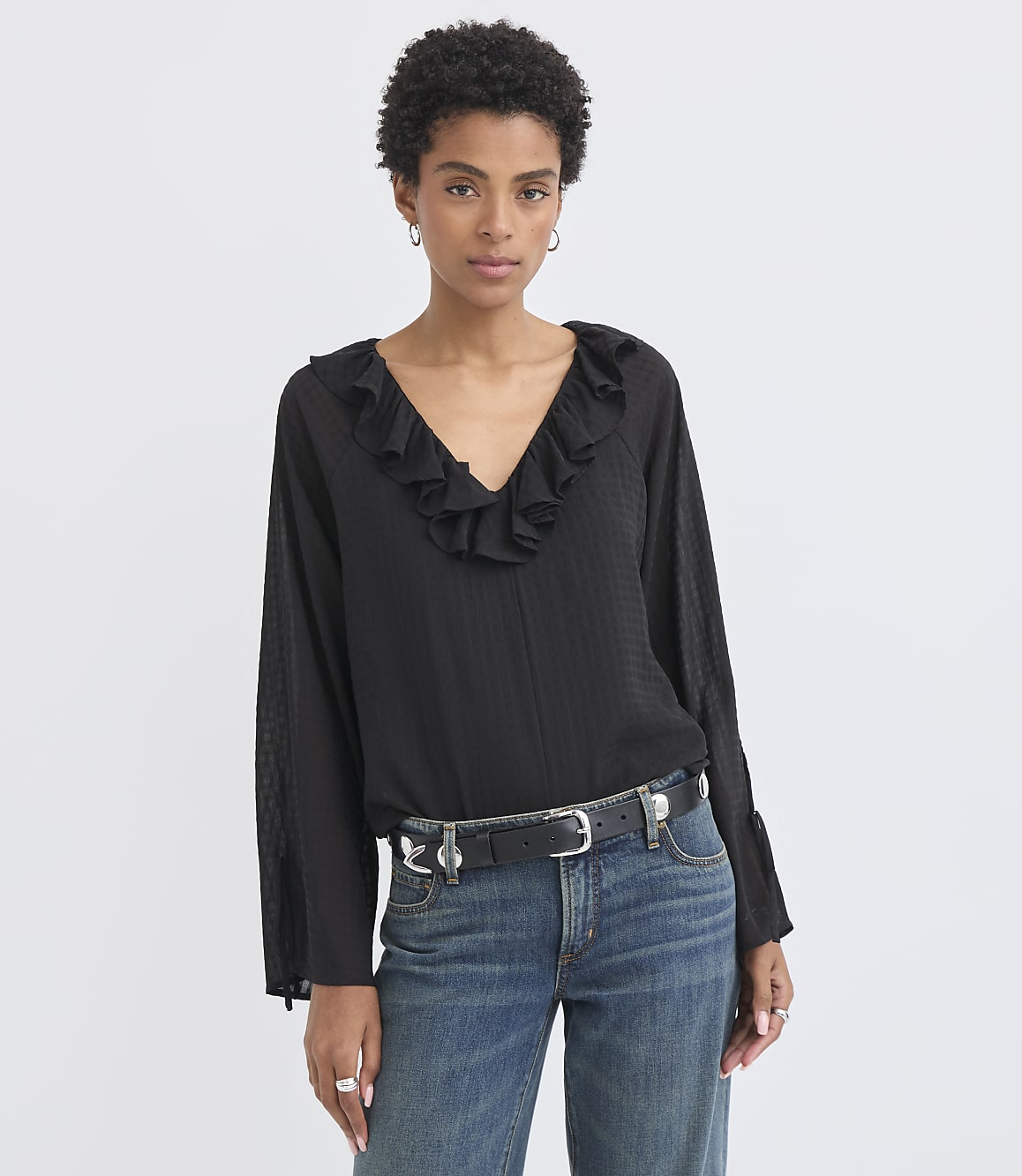 A woman in a black ruffled blouse and jeans stands against a plain white background.
