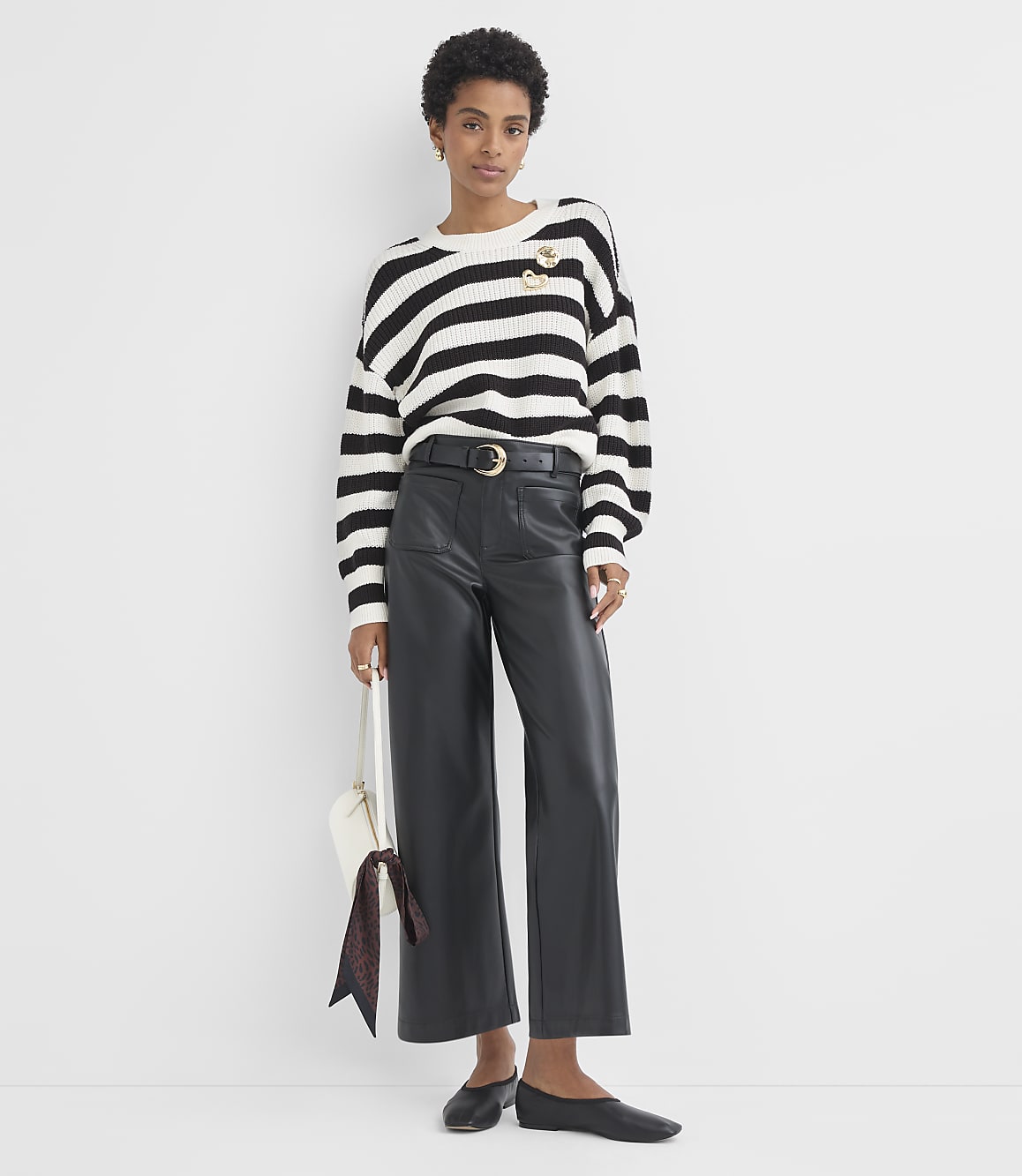 A woman in a striped sweater and leather pants stands against a plain white background, holding a white purse with a patterned scarf.