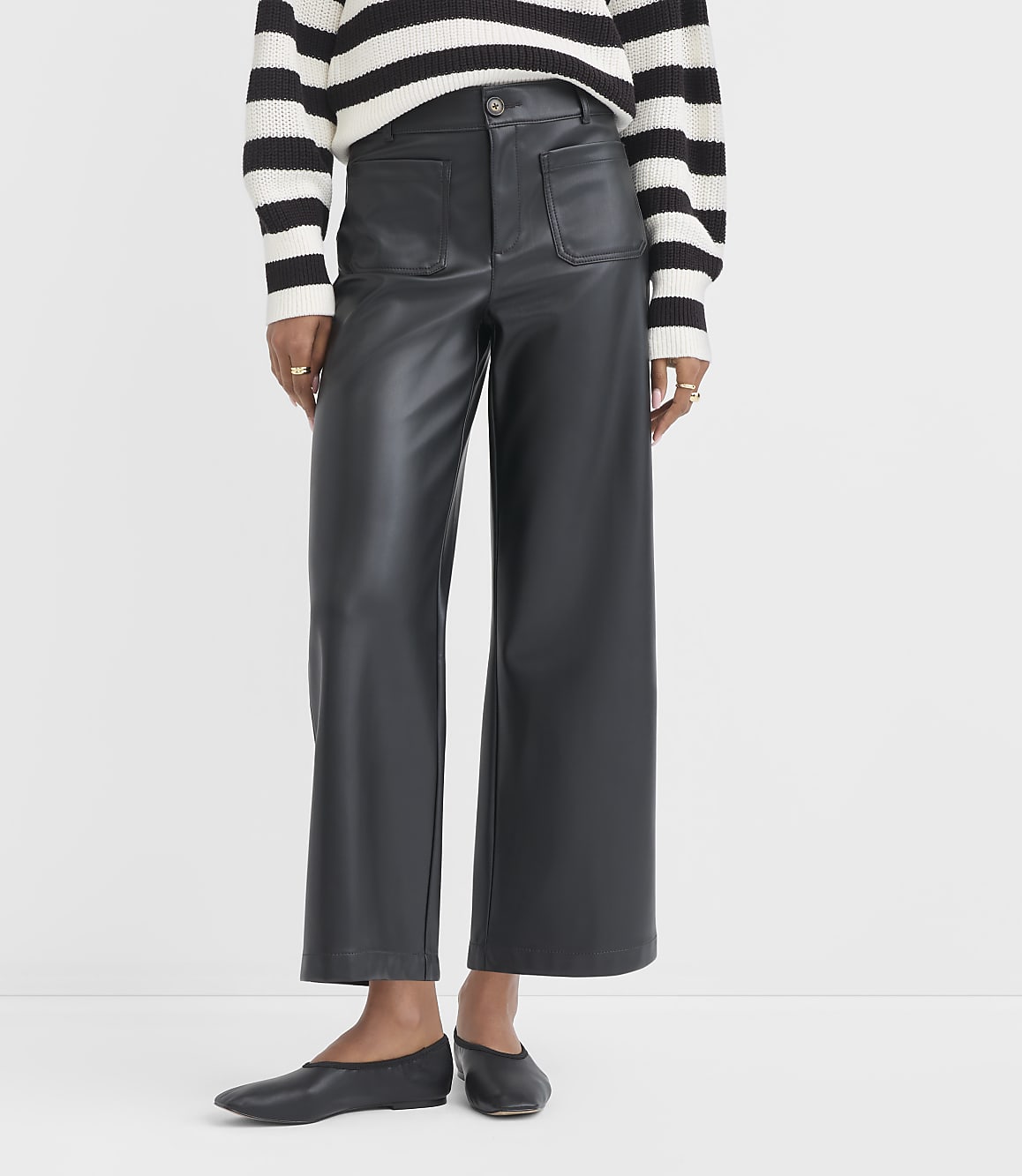 A person wearing black faux leather pants and a striped sweater stands against a plain white background.