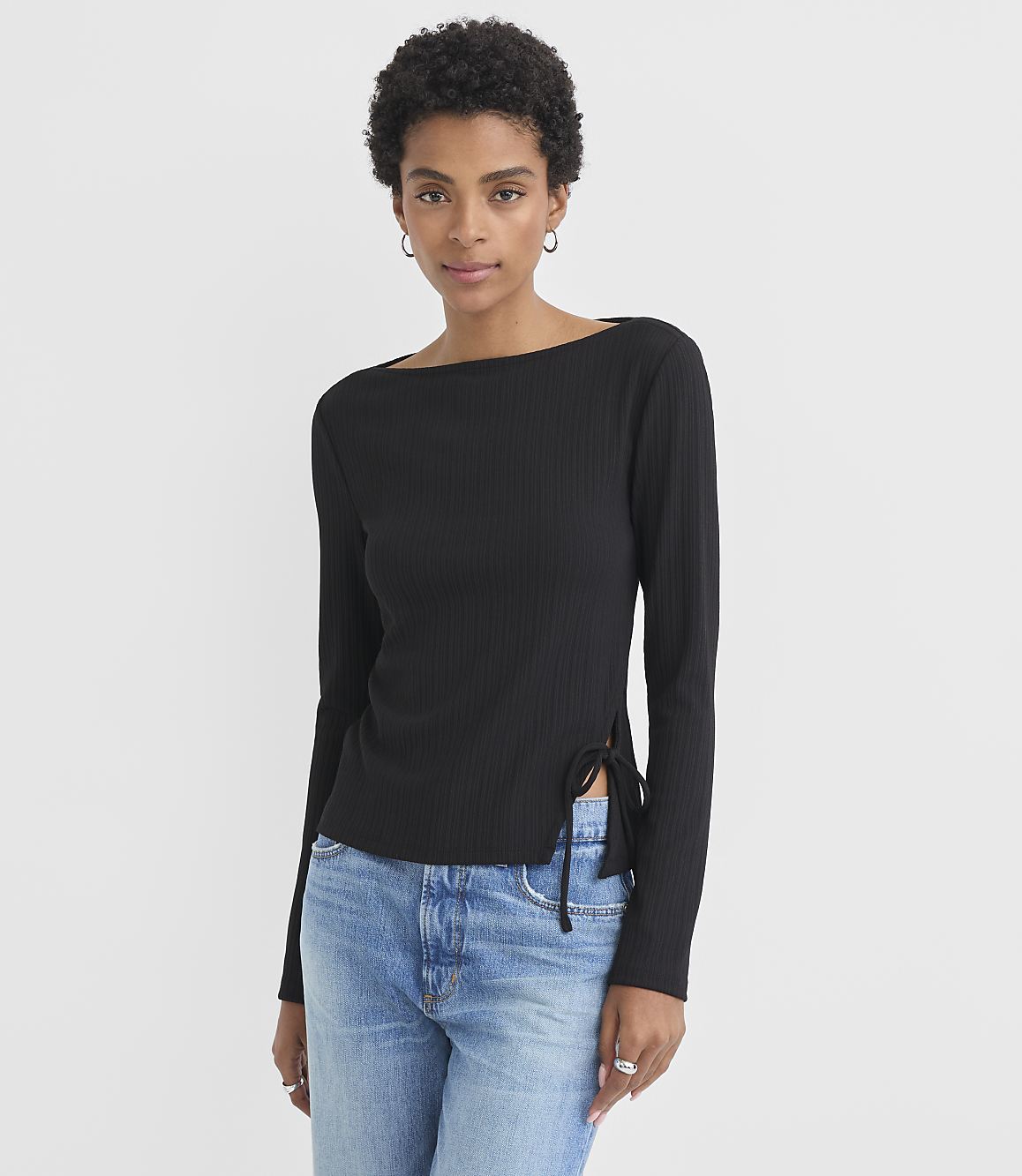 A woman in a black ribbed top and blue jeans stands against a plain white background.