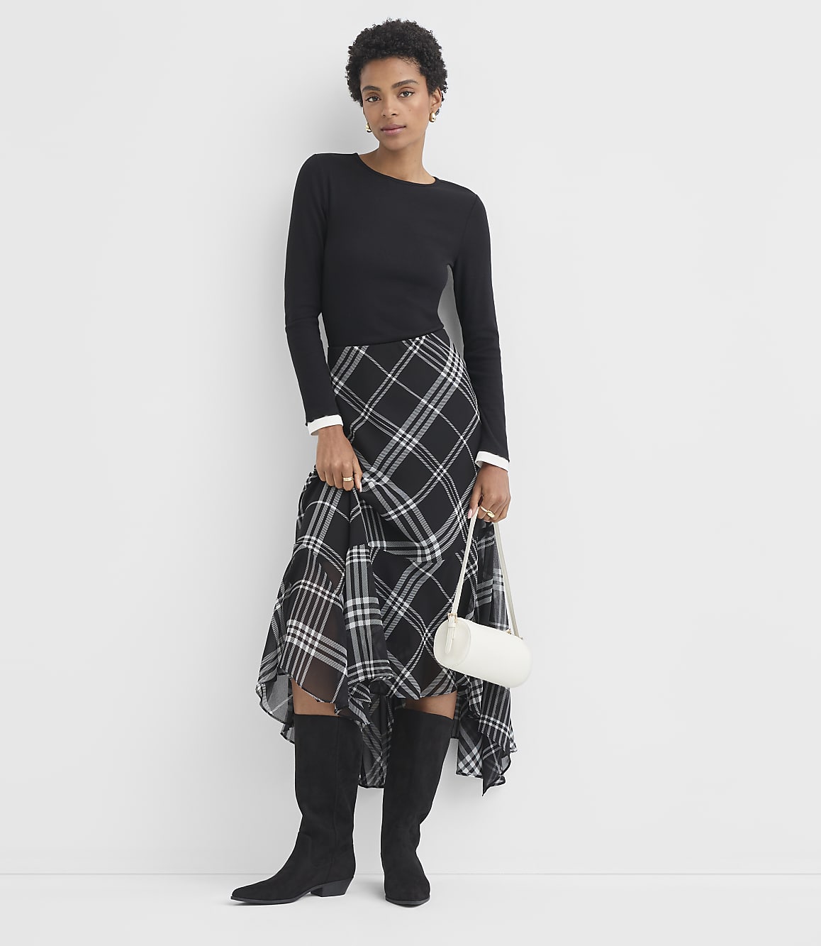 A woman in a black long-sleeved shirt and a black and white plaid skirt stands against a plain white background, holding a small white purse.