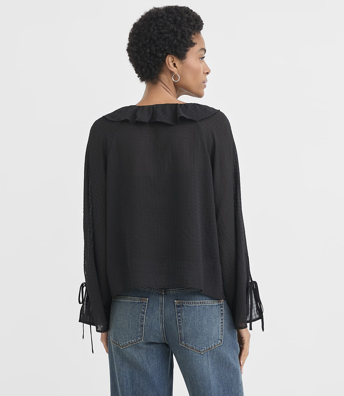 A woman in a black ruffled blouse and blue jeans stands against a plain white background.